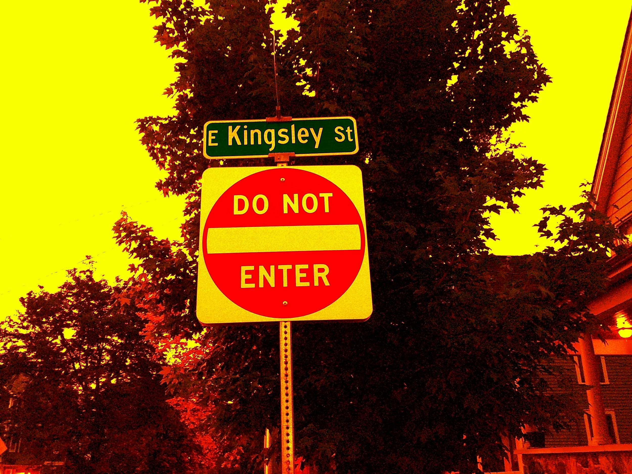 A street sign showing 'E Kingsley St' above a 'Do Not Enter' sign, set against a background of trees and building. The scene appears to be during dusk with yellow-orange lighting.