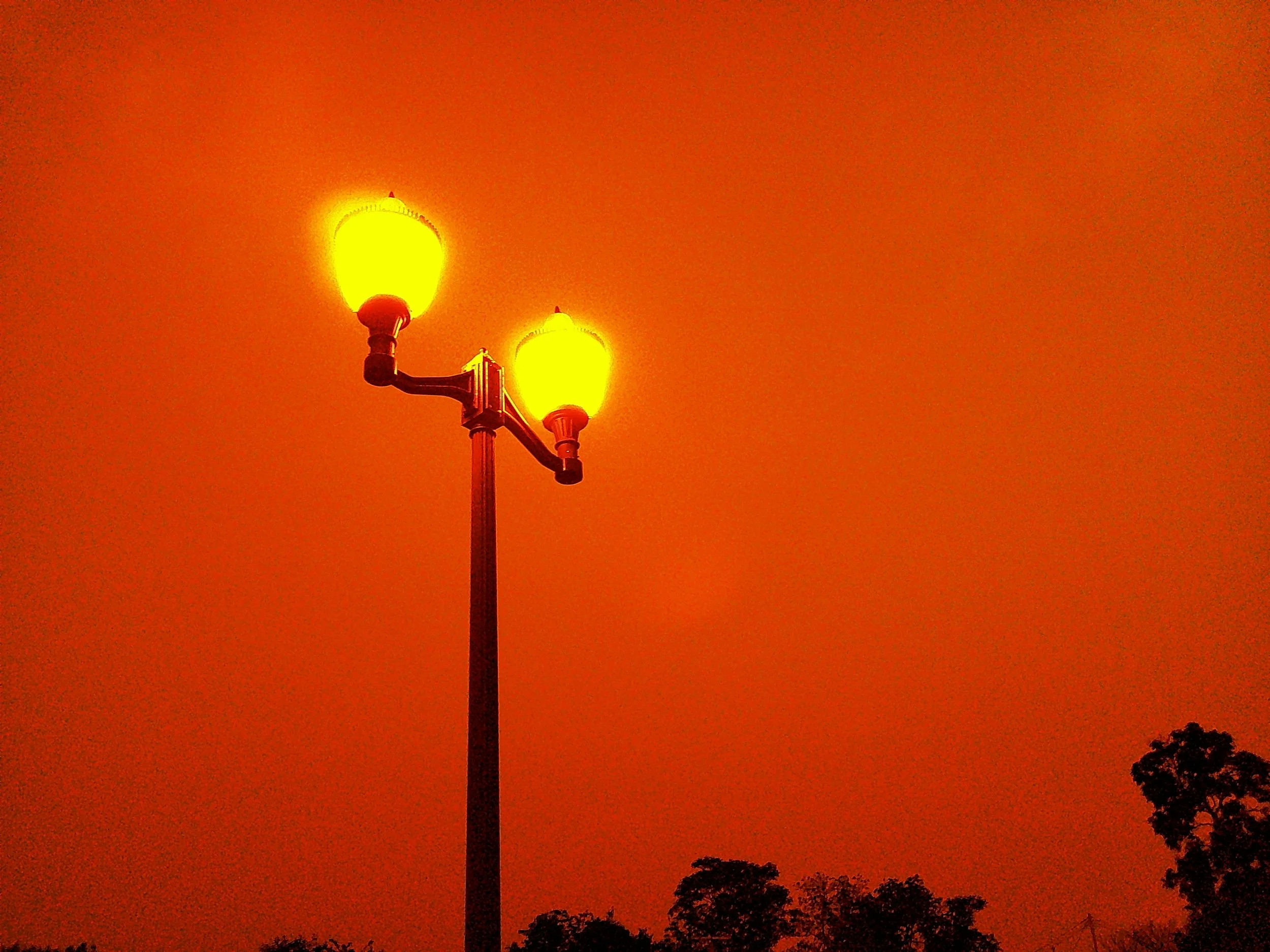 A streetlamp with two glowing yellow-orange lights against a dark orange sky, with silhouettes of trees at the bottom.