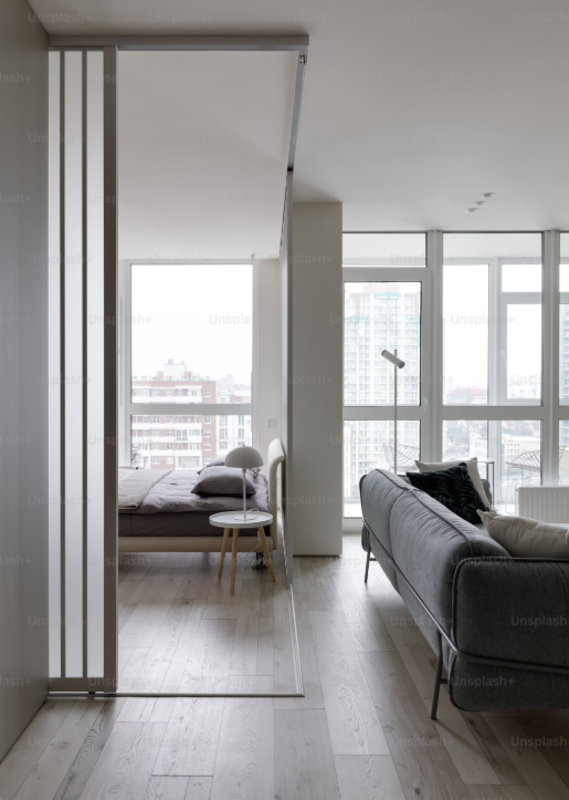 Modern apartment interior with a living room and bedroom separated by a sliding glass door, large windows with city views, gray sofa, bed with pillows, side table, lamp, and hardwood floors.