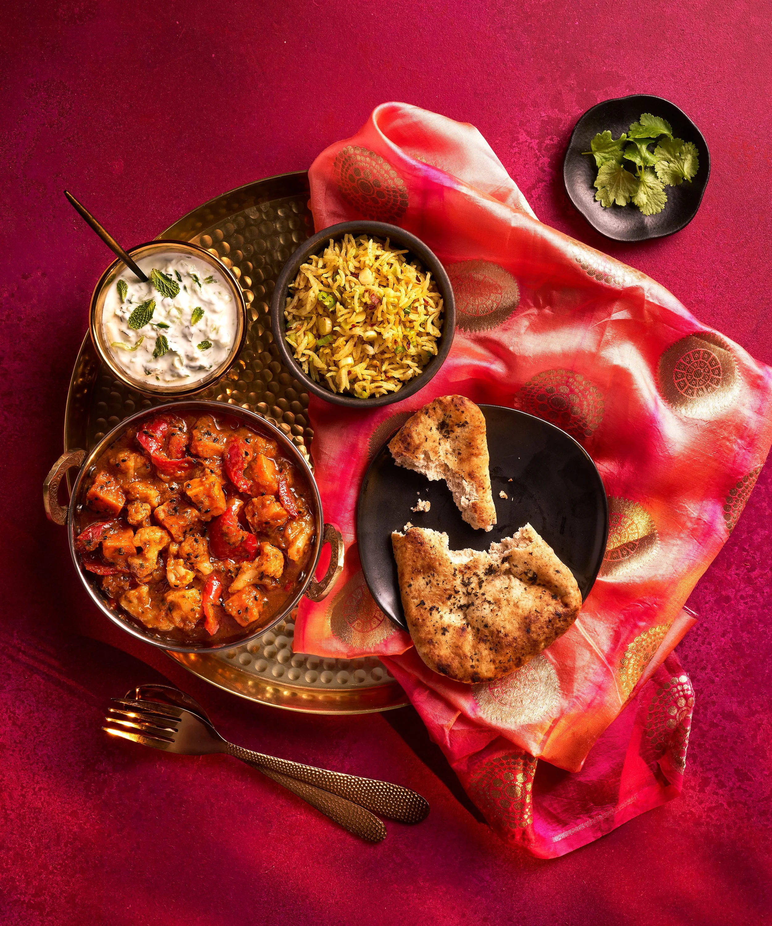 Indian meal with rice, curry, naan bread, raita, and cilantro on a brass tray with a red cloth.