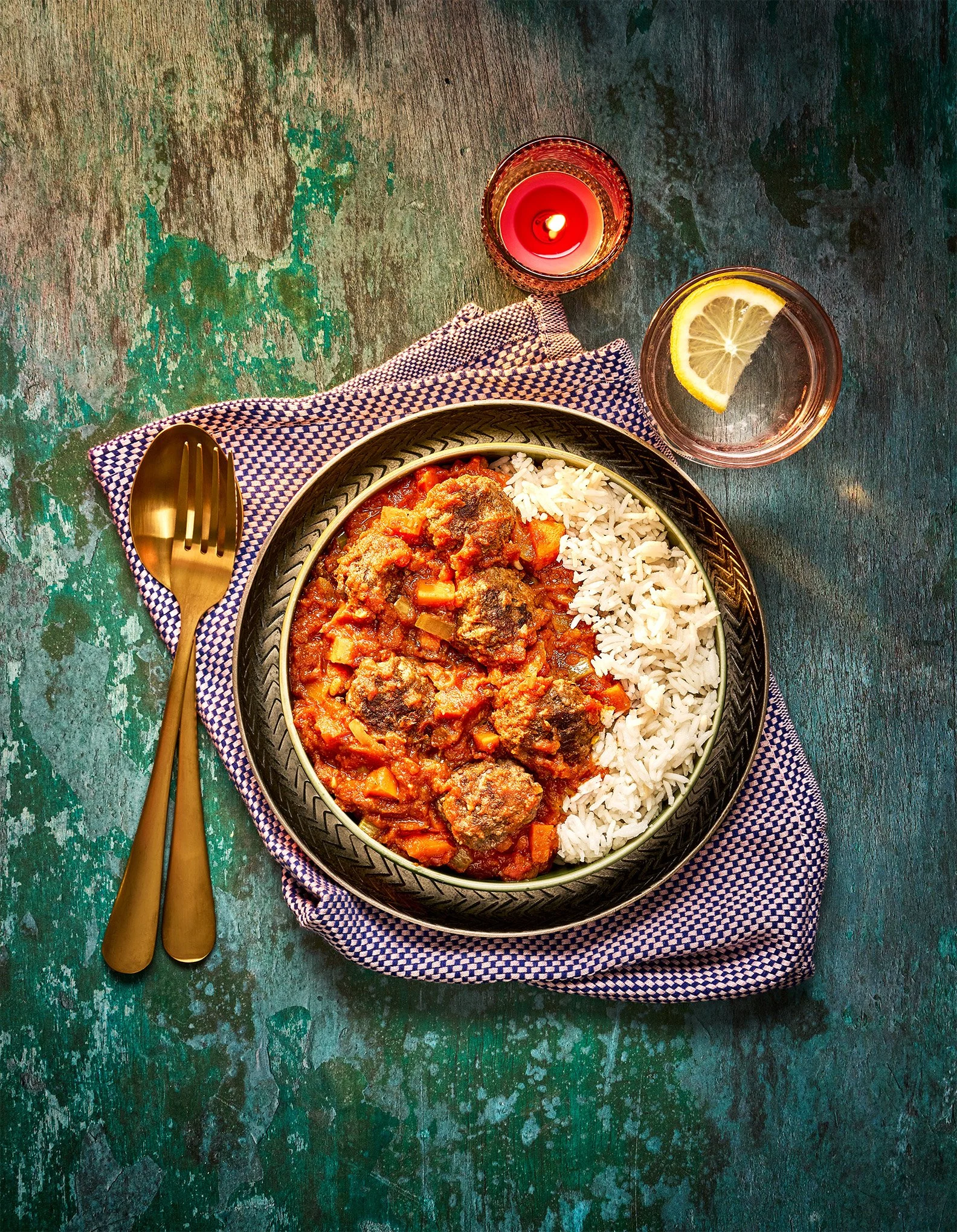 A bowl of beef stew with rice on a patterned plate, placed on a blue and white cloth napkin. A glass of water with a lemon slice, a small red candle, and a gold fork and spoon set are also on the wooden surface.
