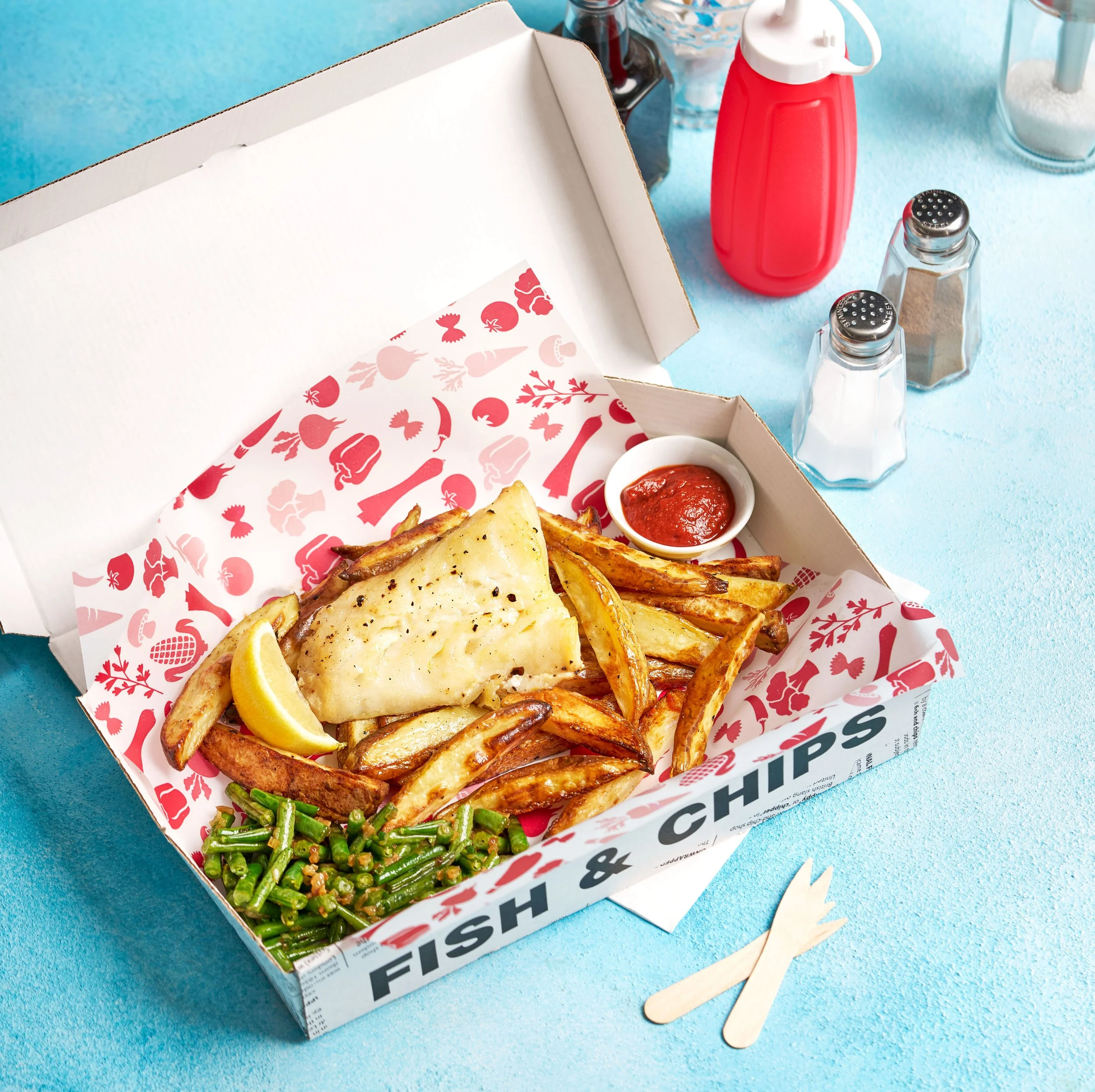 A takeout box of fried fish and chips with a side of green beans and a lemon wedge, along with a small bowl of ketchup, on a blue surface with condiments and utensils.