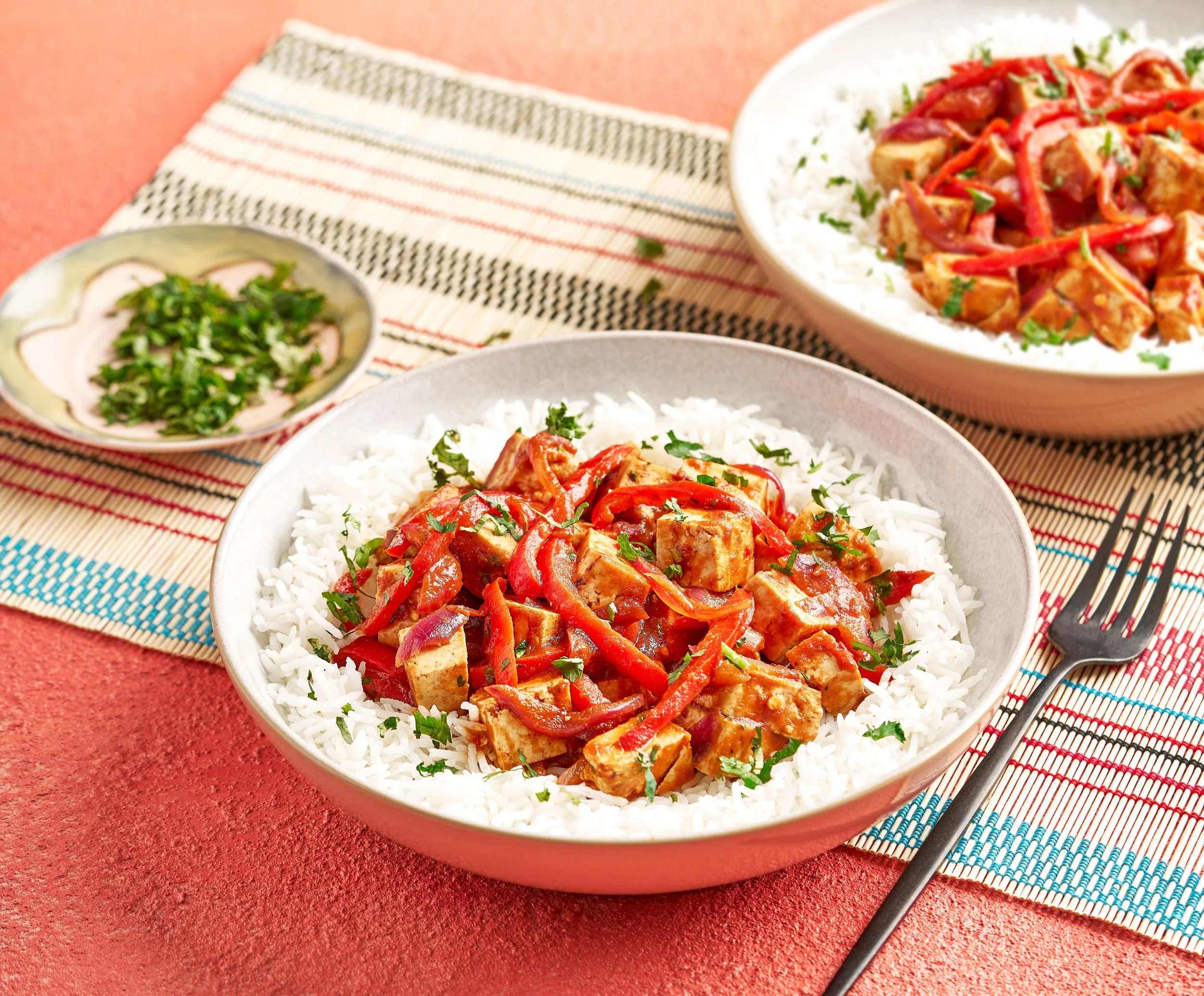 Bowls of rice topped with tofu and red bell pepper stir-fry, garnished with chopped green herbs, on a colorful striped place mat, with a small dish of chopped green onions and a fork.