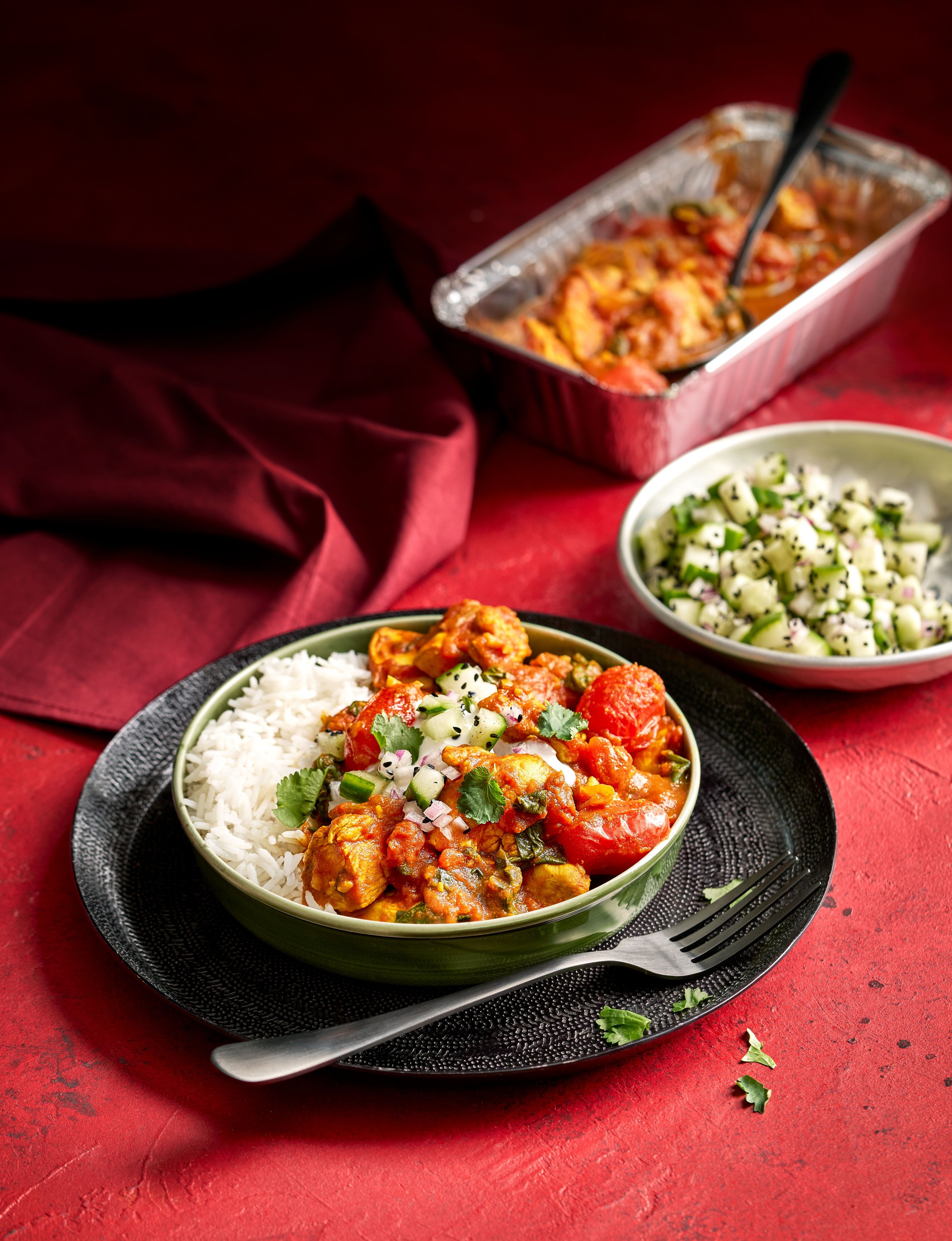 A green bowl filled with rice and vegetable curry garnished with cilantro and chopped onions, set on a black plate with a fork. In the background, there is a small bowl of chopped cucumbers and a foil tray of curry with a spoon, all on a red textured