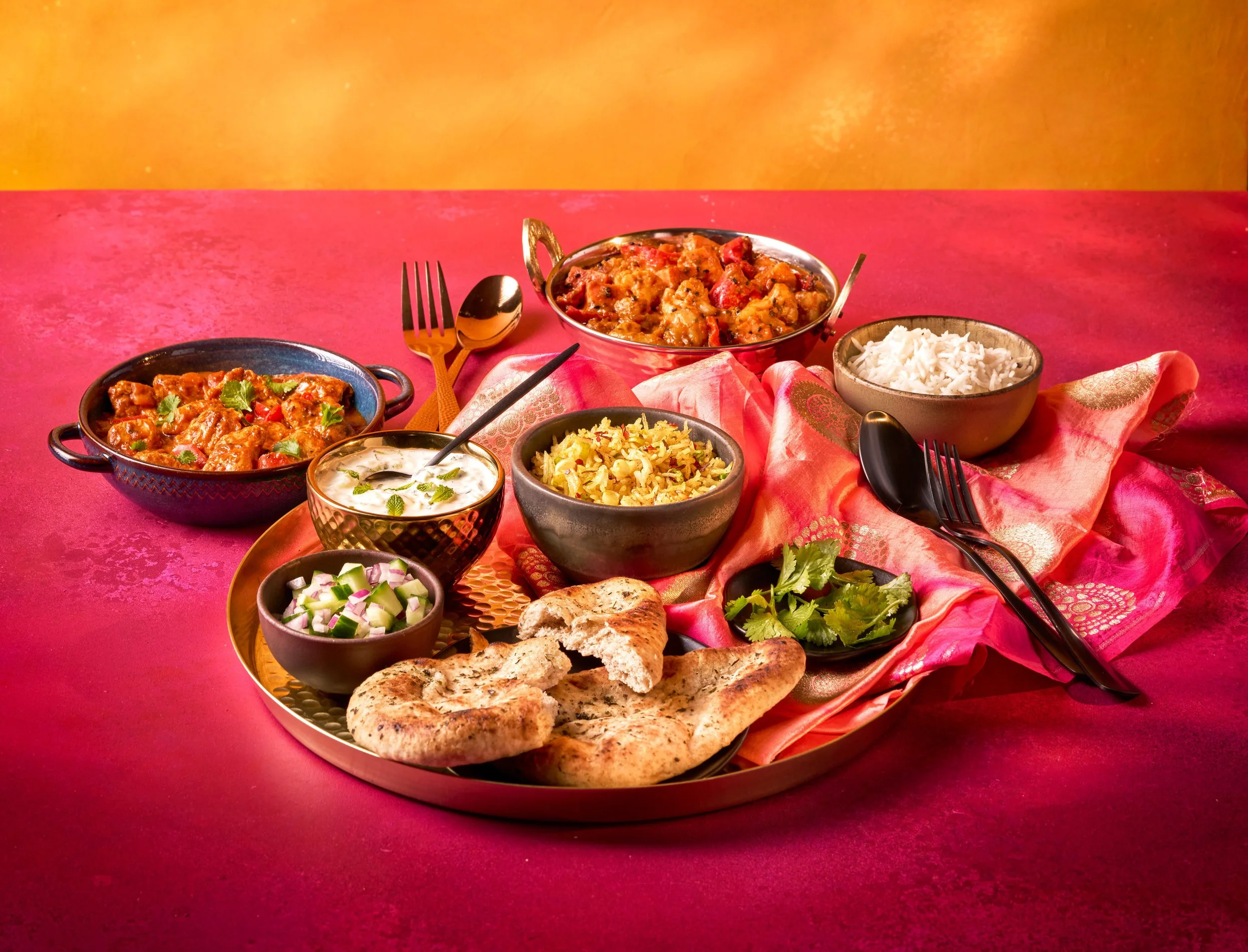 Assorted Indian dishes on a round tray with naan bread, bowls of rice, curry, raita, and vegetable salads on a pink tablecloth with a pink and gold napkin.