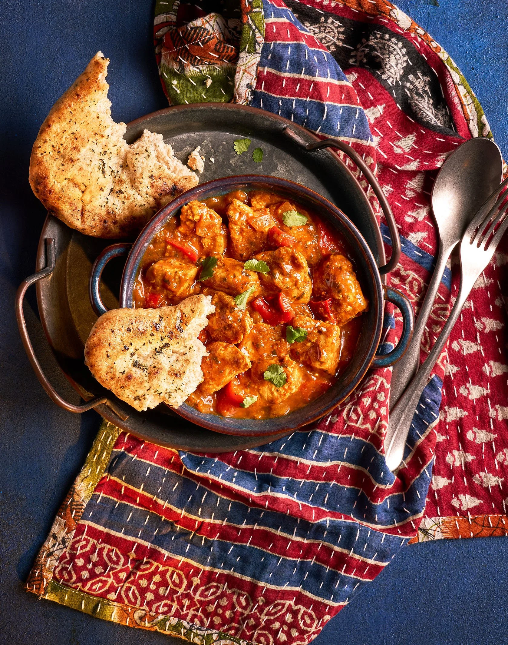 A bowl of spicy chicken curry garnished with cilantro, accompanied by toasted naan bread on a metal tray, placed on a colorful fabric with a fork and spoon nearby.