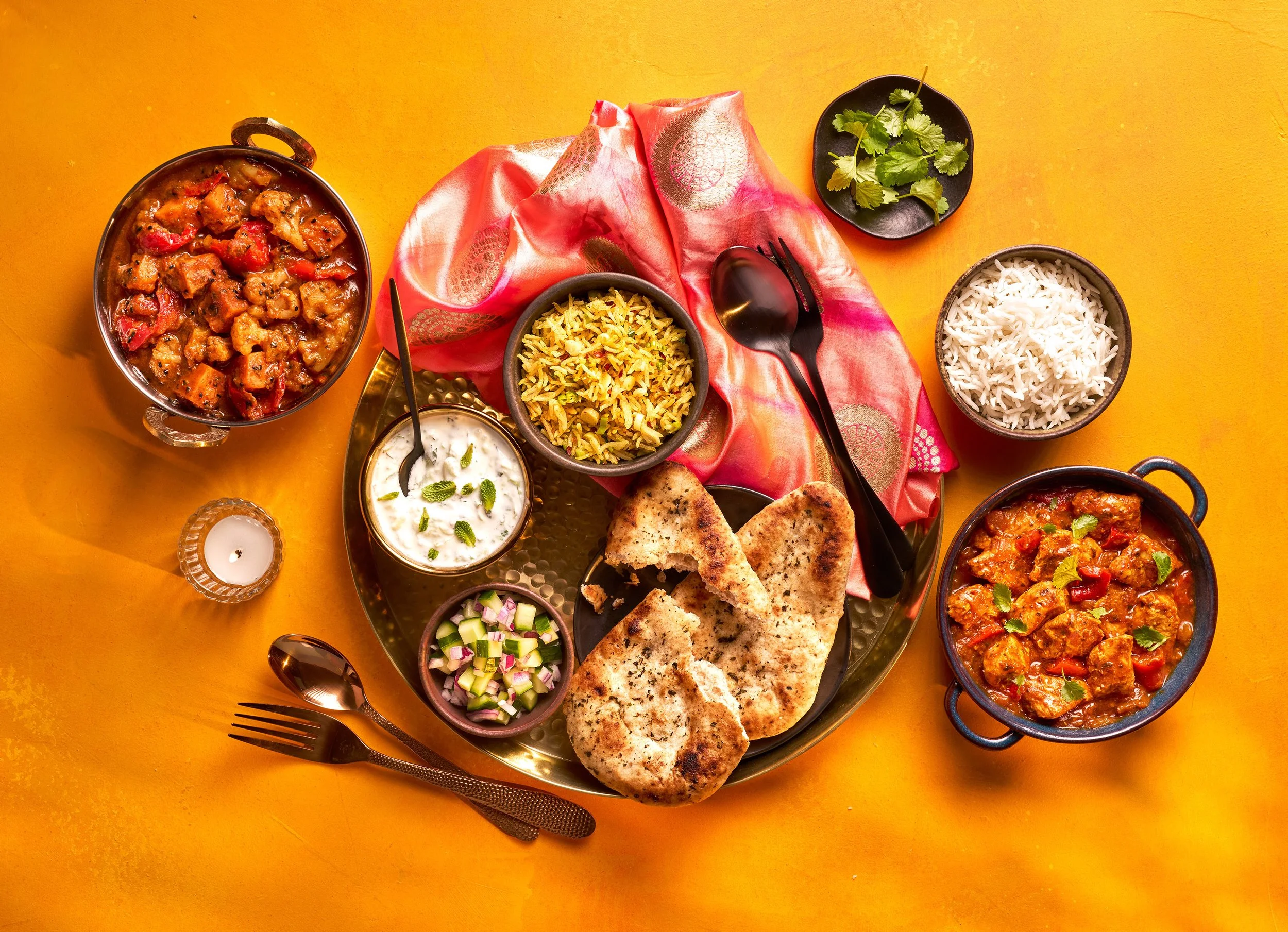 An Indian meal served on a large round tray with naan bread, rice, curry, yogurt, and garnishes, set on a yellow table with a pink cloth, silver cutlery, and a small votive candle.