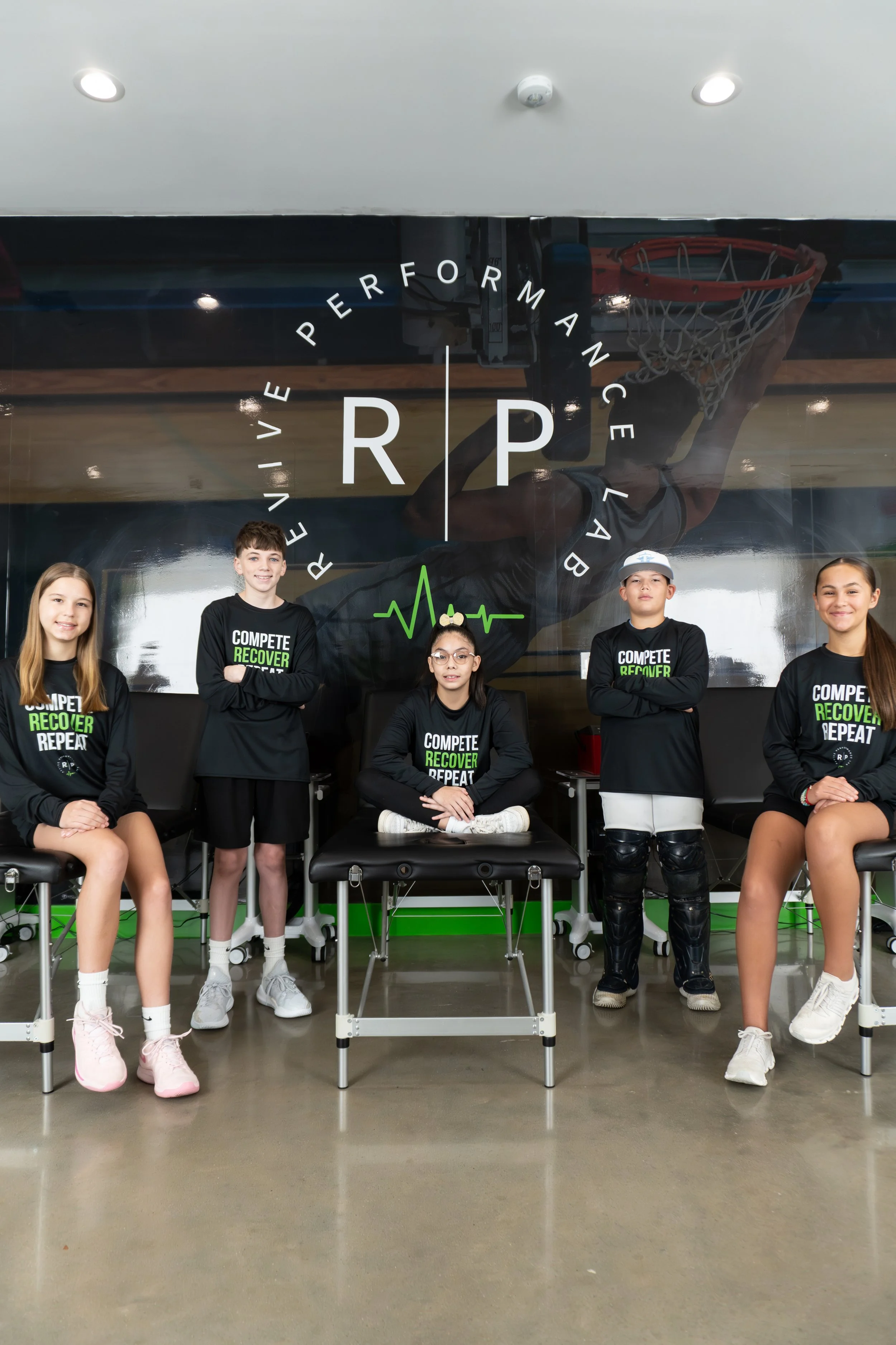 Five young athletes in black shirts with the slogan "Compete, Recover, Repeat" standing and sitting in front of a fitness or sports facility with a logo featuring a basketball hoop and the text "REVIVE PERFORMANCE LAB" visible behind them.