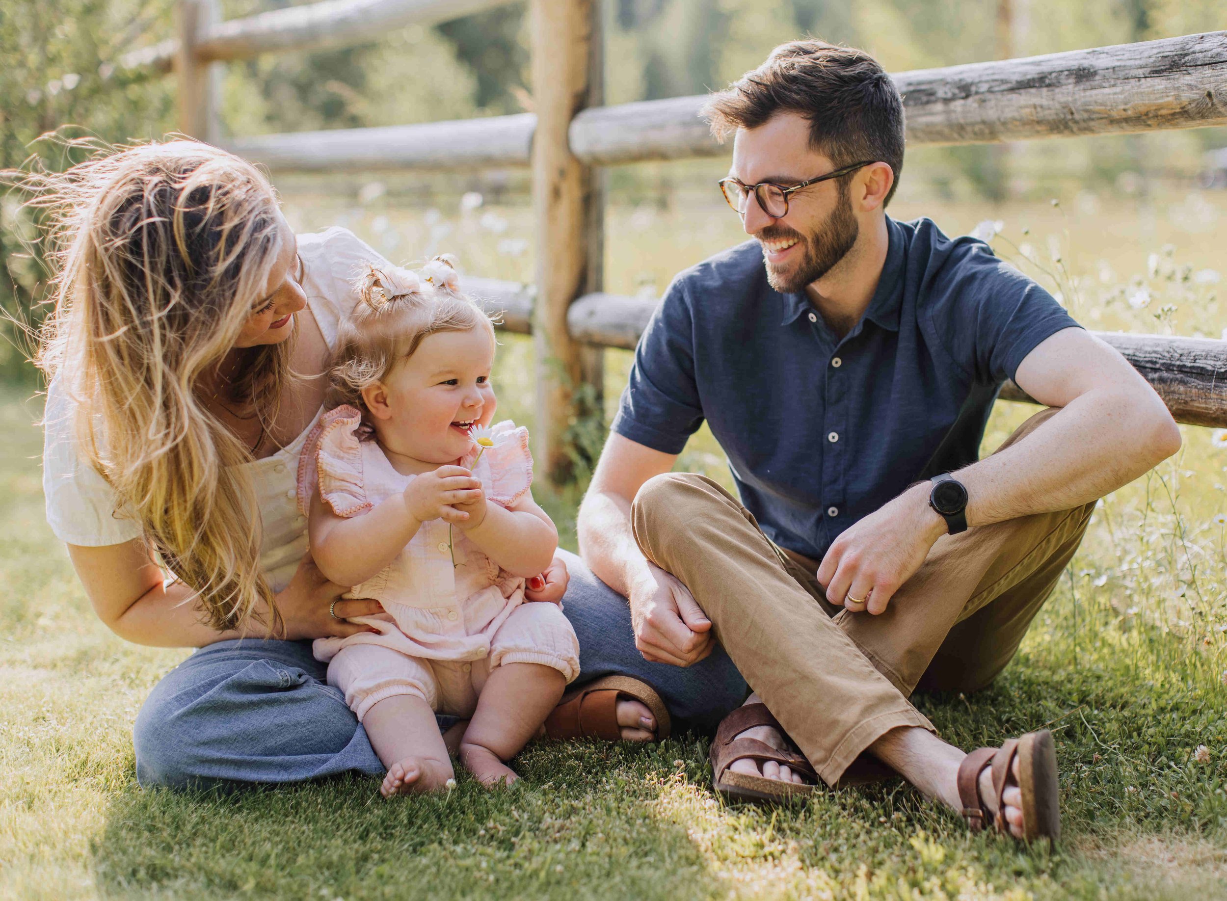 A family of three sitting on the grass outdoors, smiling and enjoying a moment together. The woman has long blonde hair, and the child is a young girl with curly hair holding a daisy. The man has short dark hair, glasses, and a beard, wearing casual