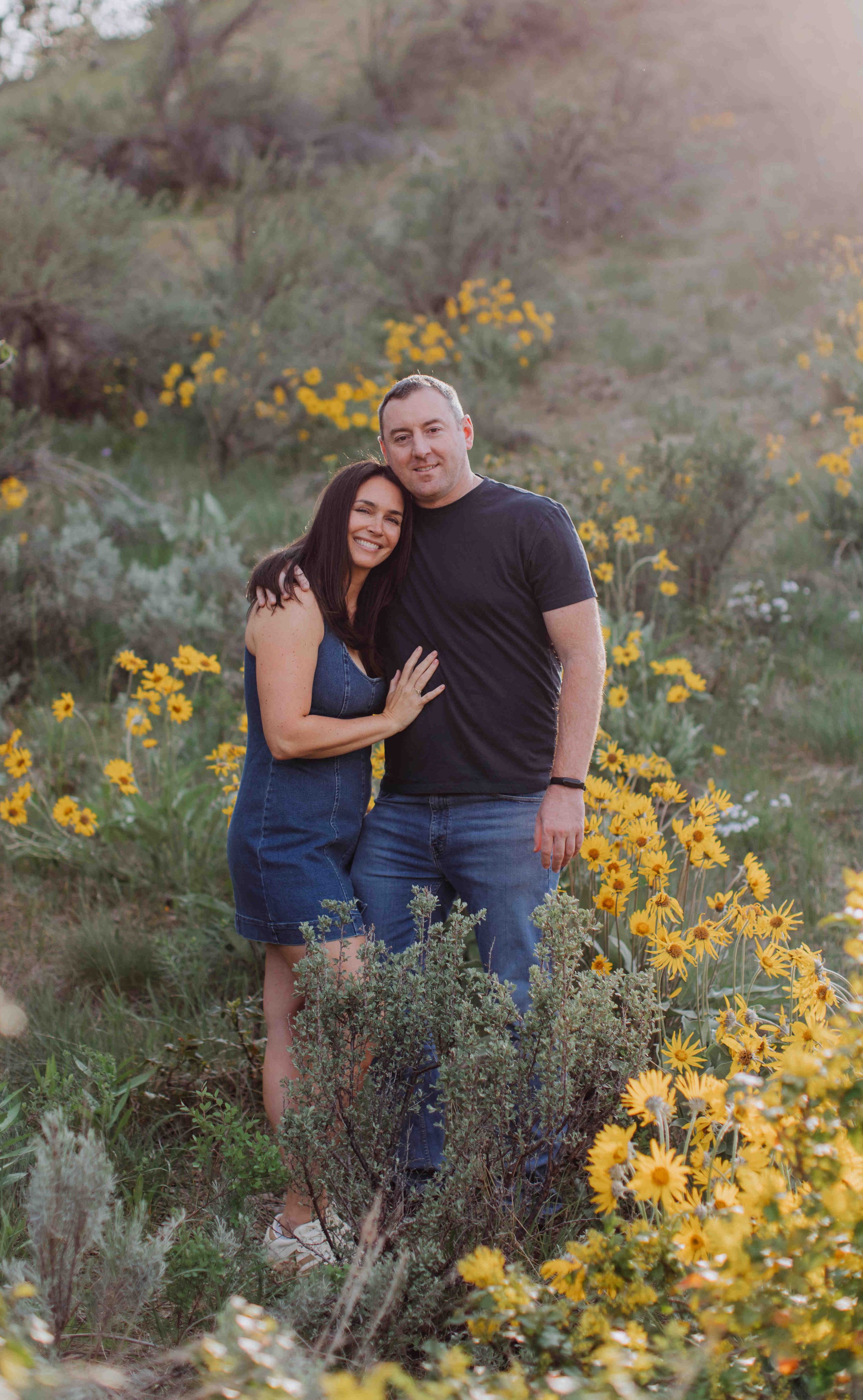 A smiling couple stands close together in a field of yellow flowers with a hilly, shrub-covered background.