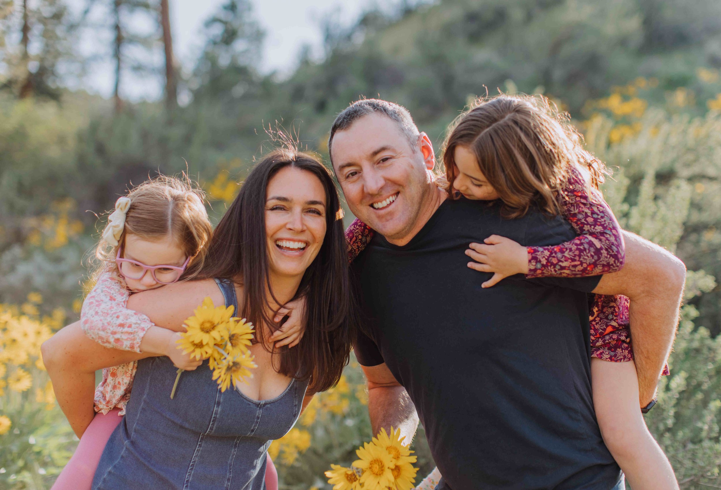 Family outdoors, two women and two children smiling and enjoying sunshine in a field of yellow flowers.