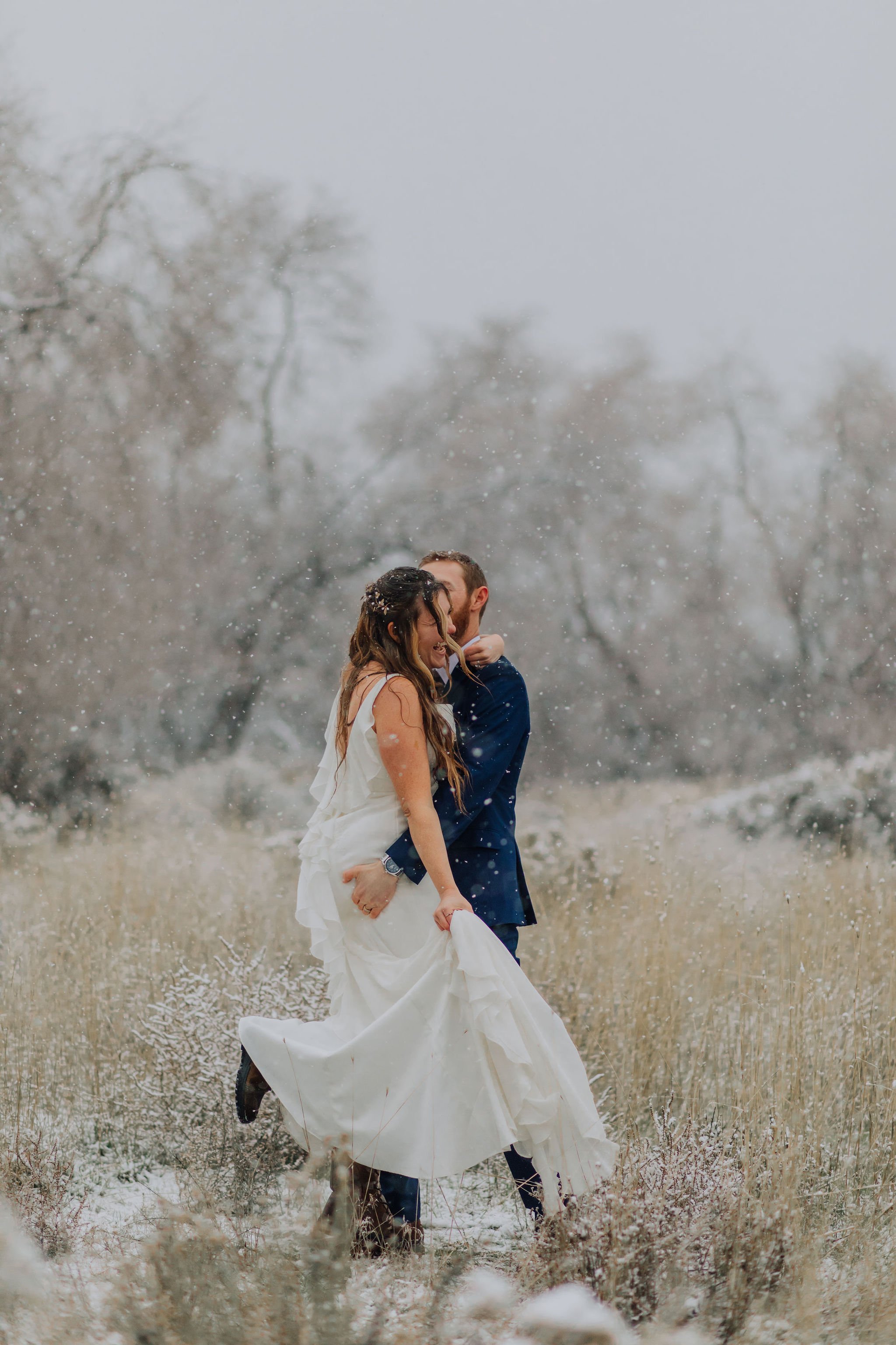 A couple in wedding attire sharing a joyful moment in a snowy outdoor setting with bare trees in the background.