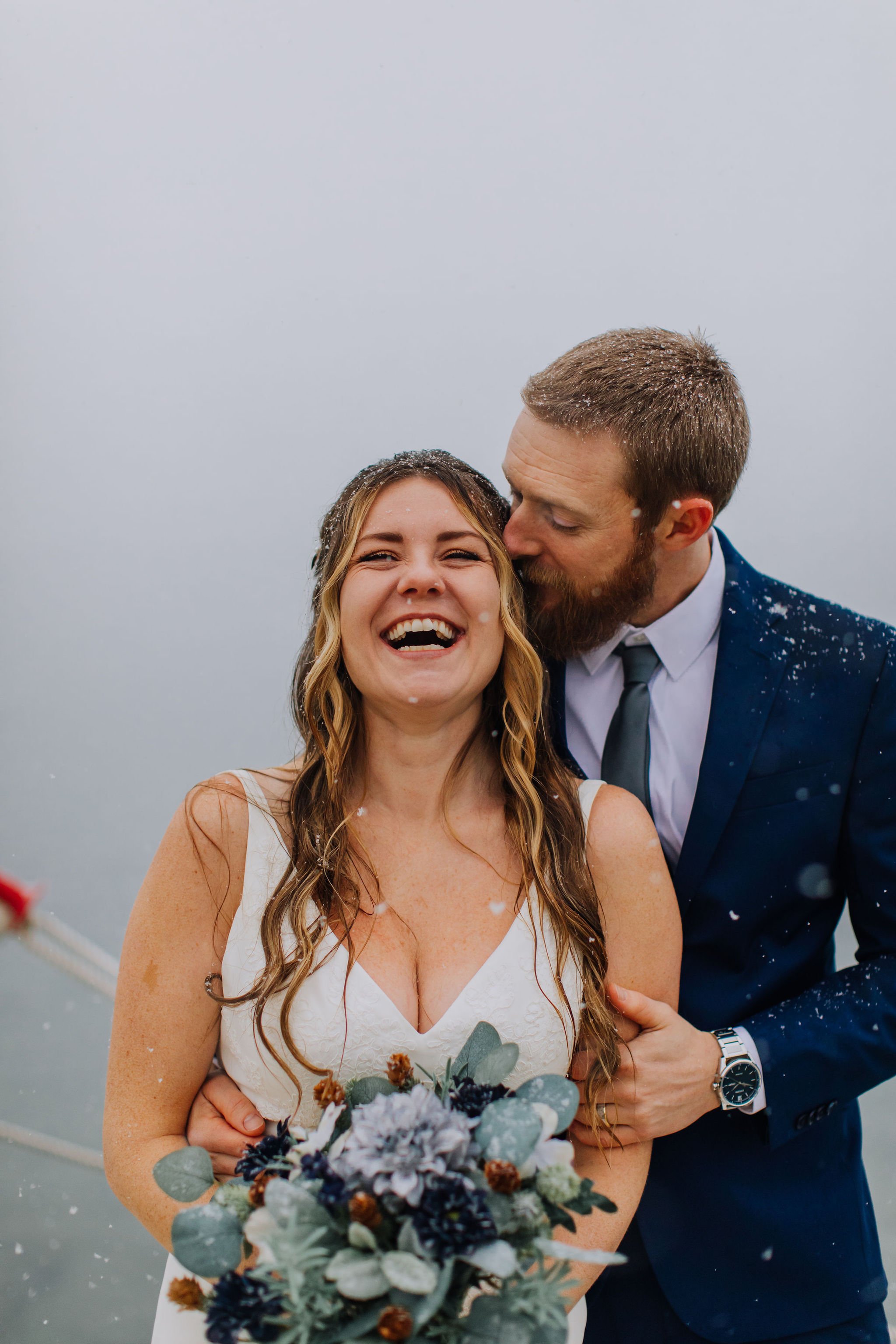 A joyful bride and groom on their wedding day, with the groom kissing the bride's temple, standing outdoors in a foggy environment, with snowflakes in the air. The bride is holding a bouquet of flowers and wearing a white dress, while the groom is dr