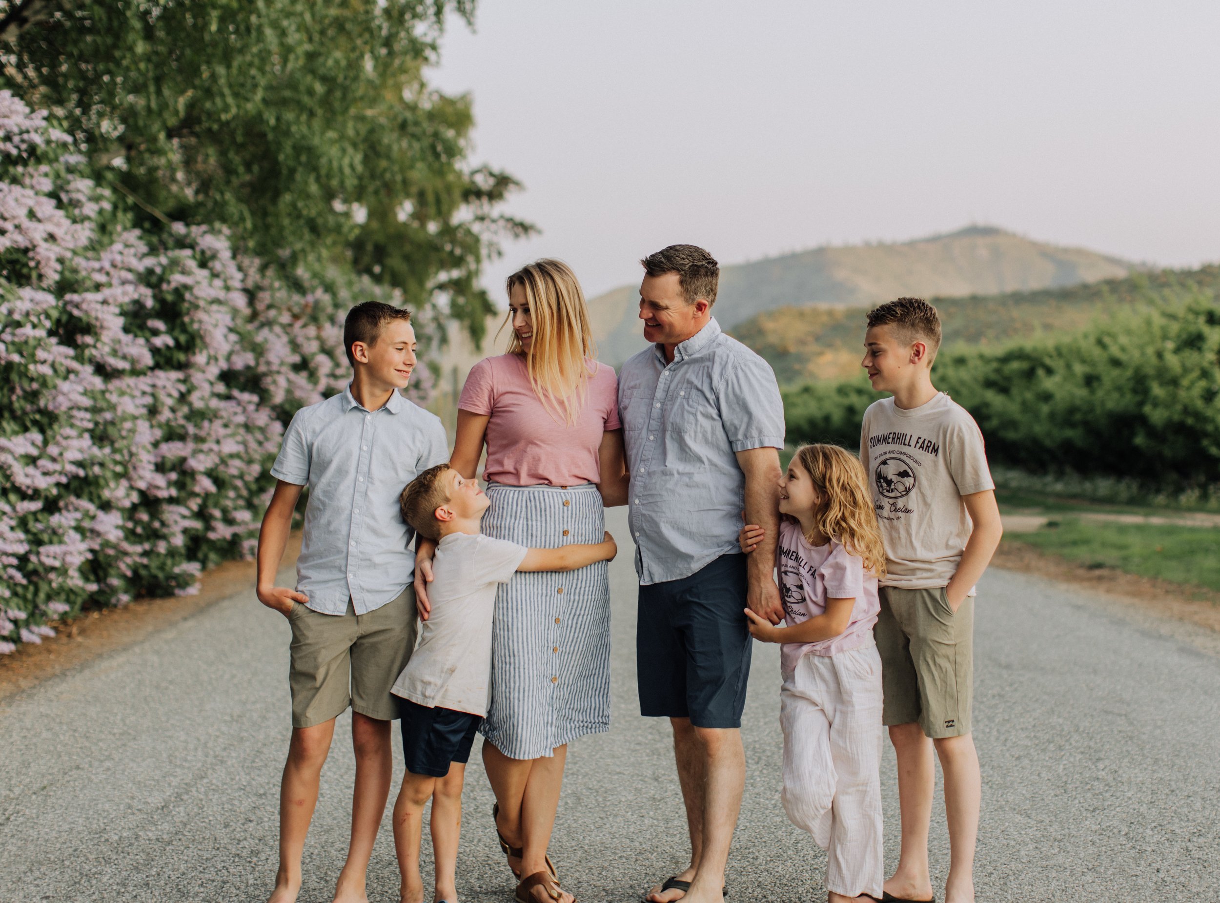 A family of seven, including a mother, father, three boys, and two girls, walking and smiling on a rural road with greenery and hills in the background, enjoying a sunny day.
