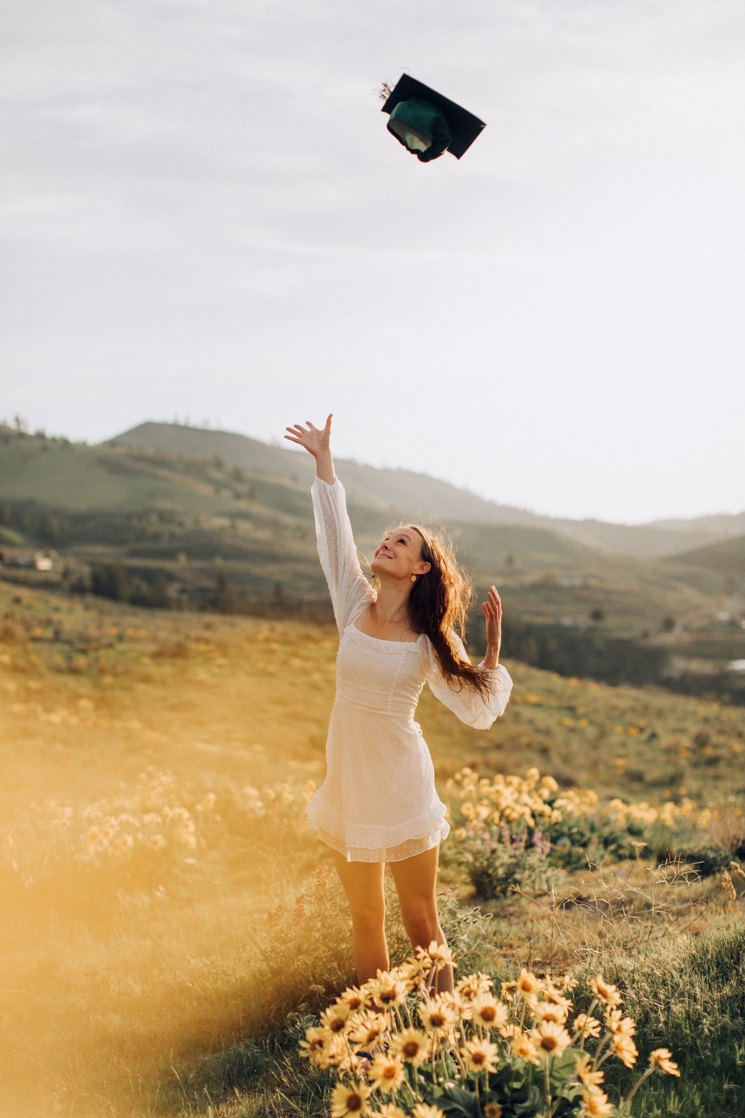 A young woman in a white dress standing in a field of yellow flowers, tossing a graduation cap into the air with hills in the background.