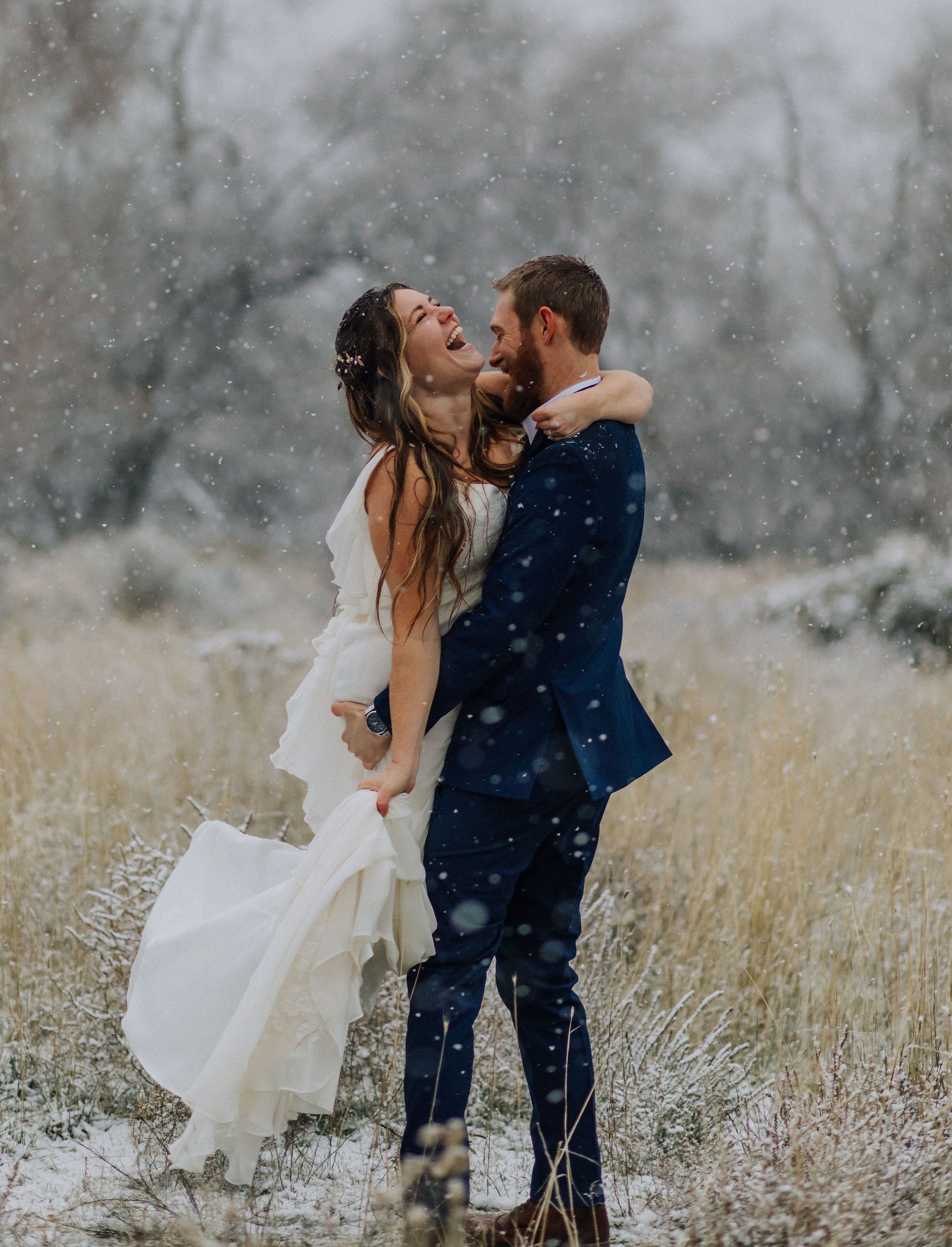 A bride and groom are outside in a snowy landscape, embracing and laughing while dancing.