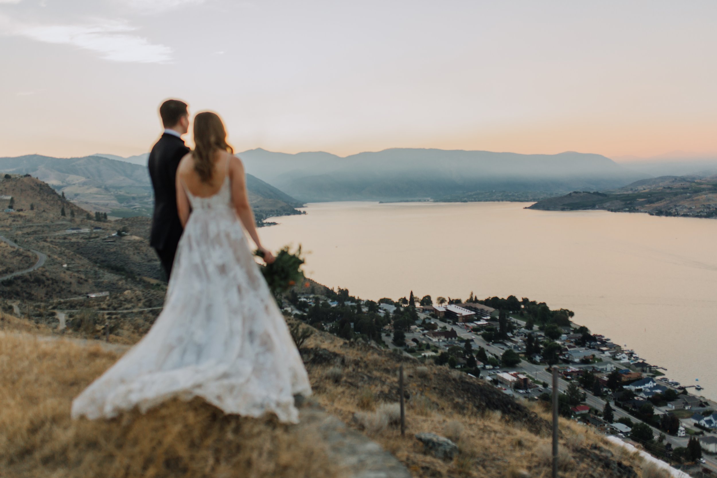 A newlywed couple stands on a hillside overlooking a lake and town at sunset, with mountains in the background.