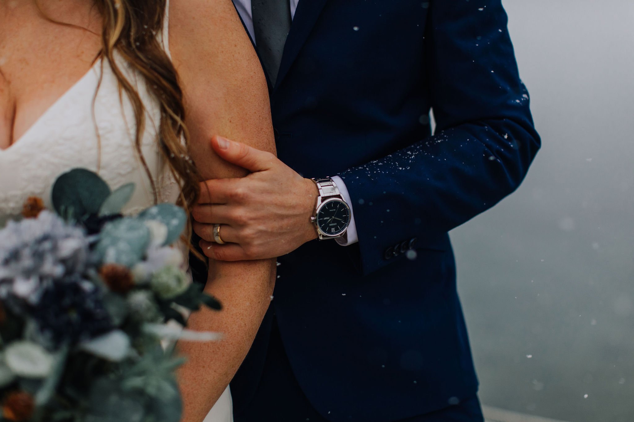 Close-up of a groom's hand with a watch holding the bride's arm during a wedding ceremony, with a bouquet in the foreground and a blurry gray background.