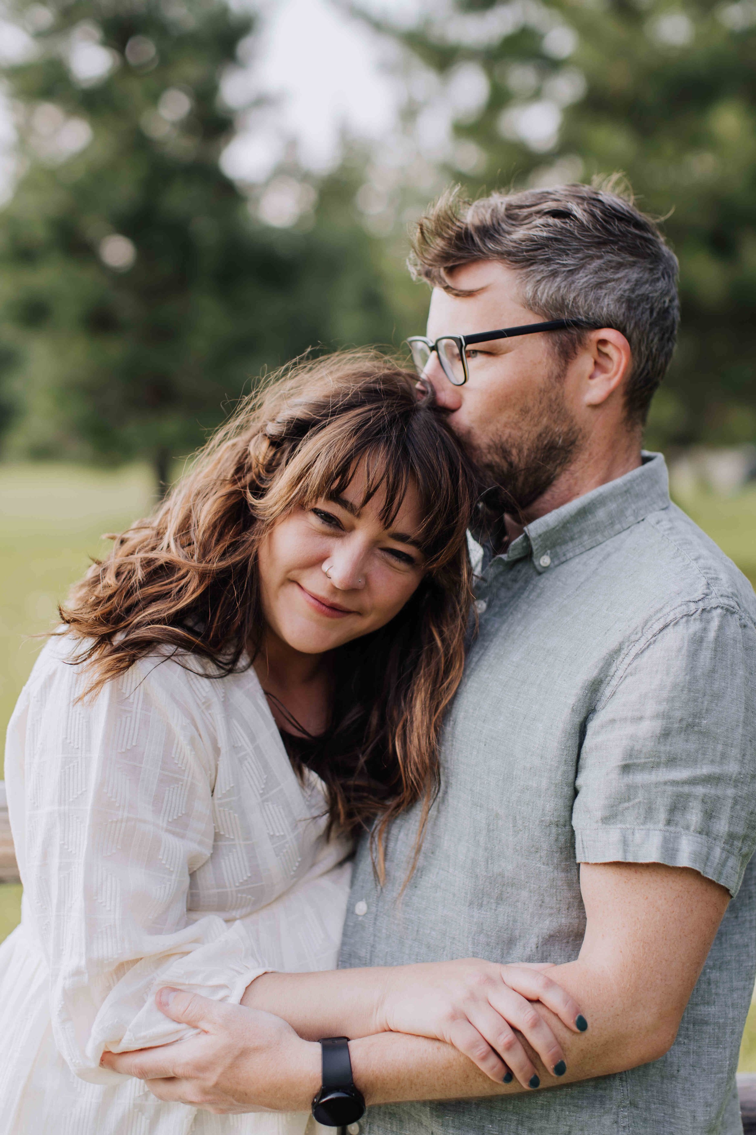 A man and woman embrace outdoors, with the man kissing the woman's forehead. The woman has brown hair with bangs, a nose ring, and is smiling softly. The man has glasses, a beard, and gray hair, and is wearing a gray button-up shirt. They are standin