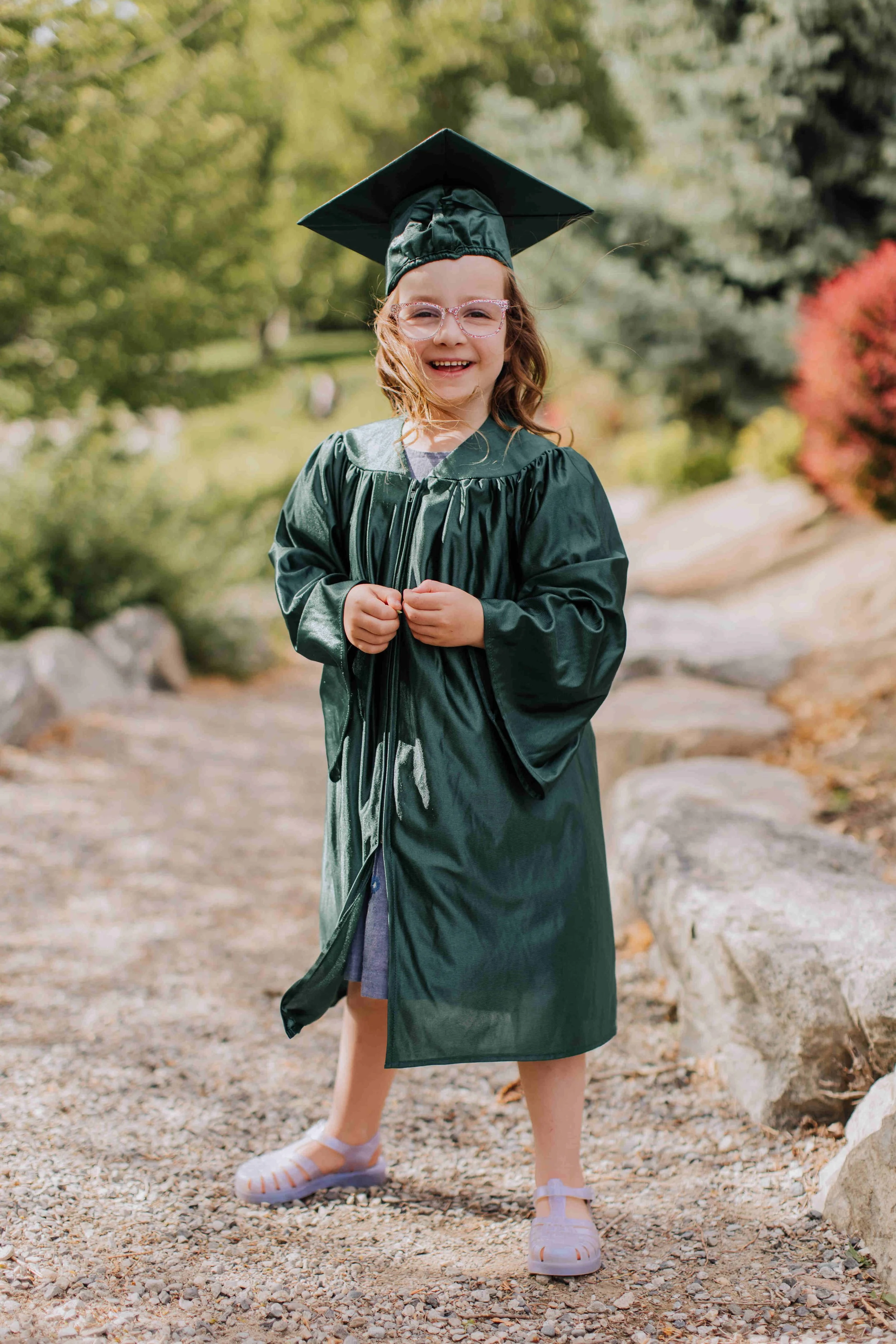 Smiling kindergarten graduate in a green cap and gown standing on a path at Riverwalk Park in Chelan during a Petite family photography session.
