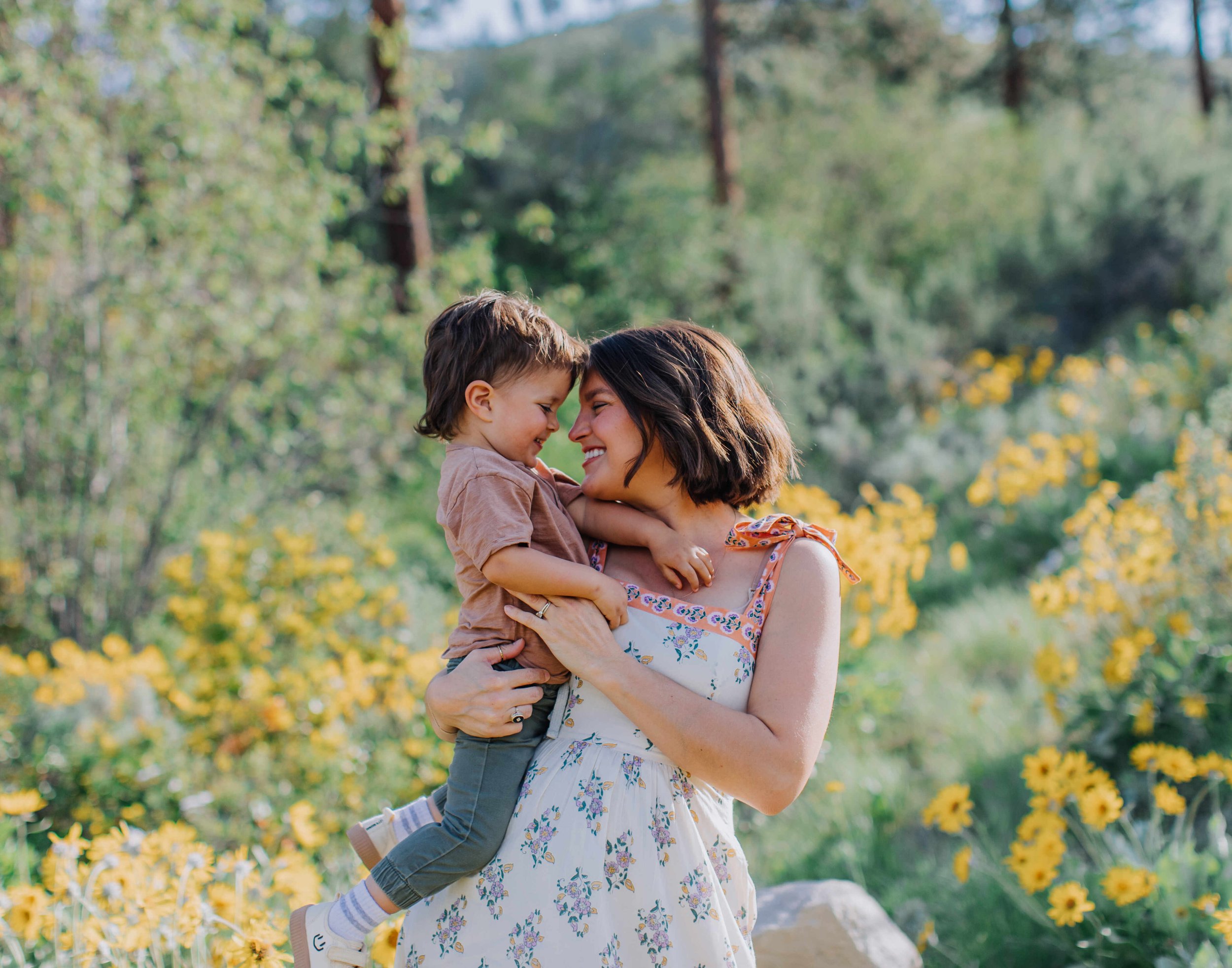A woman and young boy, likely mother and son, are outdoors in a field with yellow flowers, smiling and touching foreheads, sharing a joyful moment.
