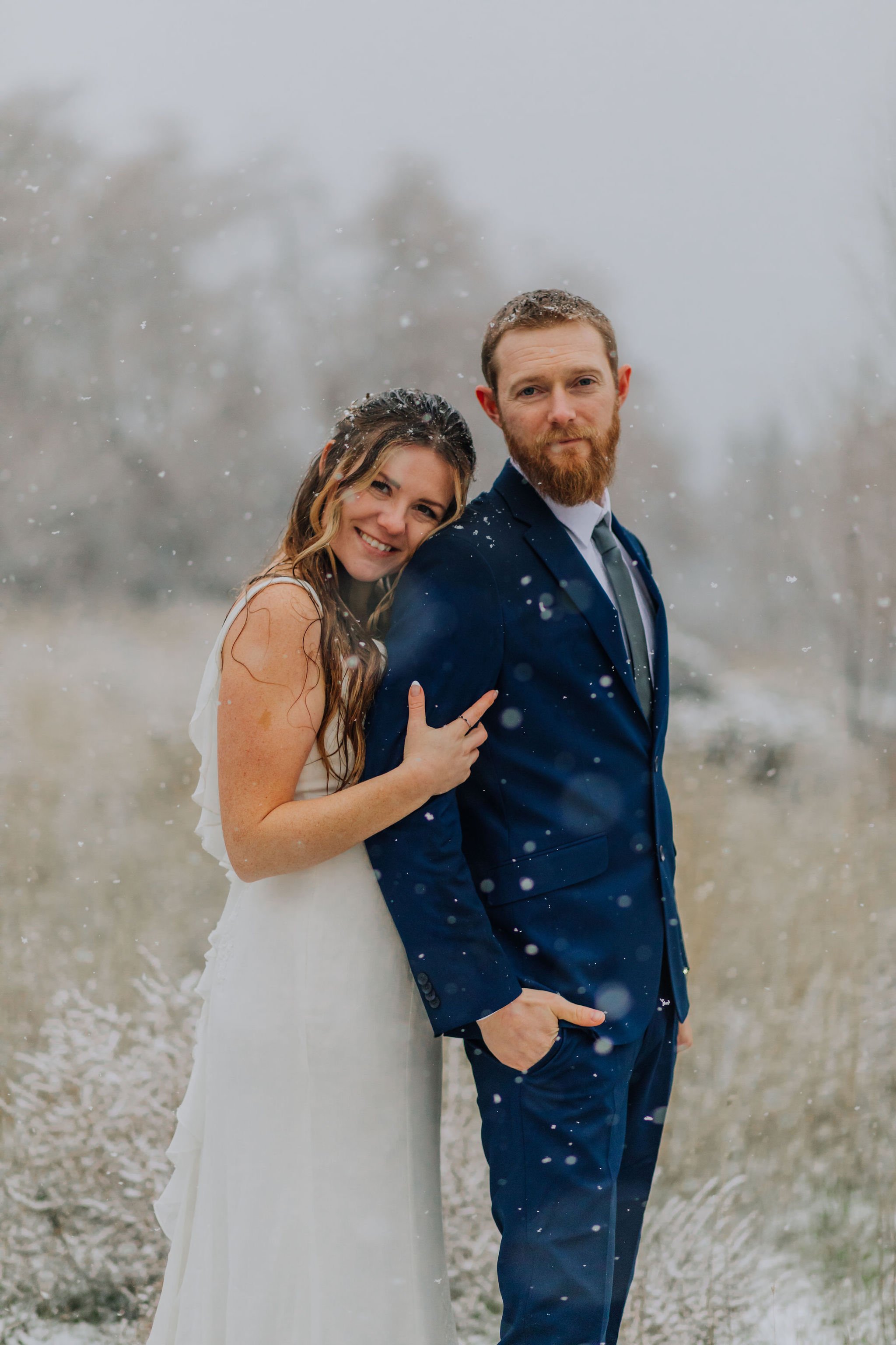 A bride and groom standing outside in a snowy landscape, with the bride leaning on the groom's shoulder and smiling at the camera.