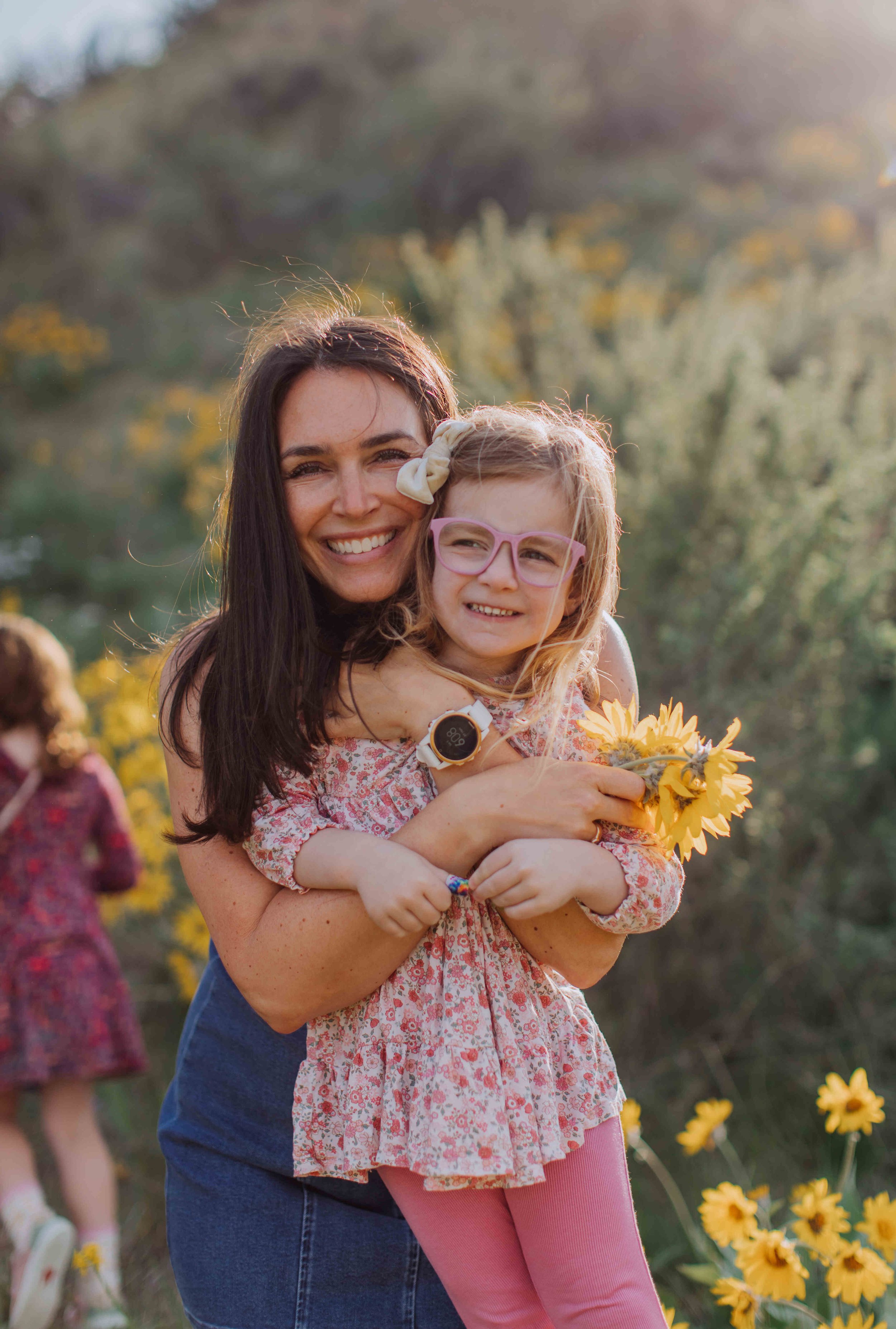 A woman hugging a young girl holding a bouquet of yellow flowers outdoors with another girl in a similar dress in the background.