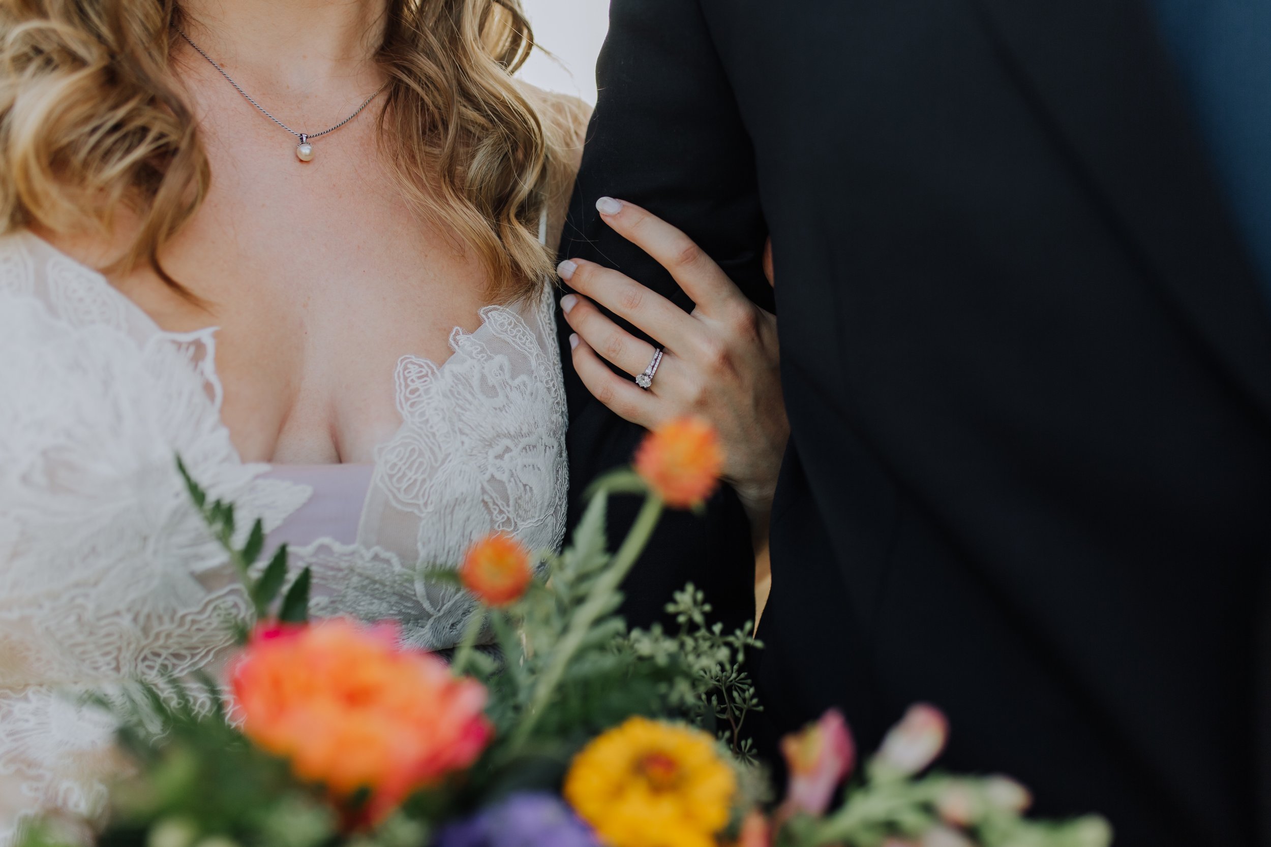 Close-up of a wedding couple showing the bride's wedding ring and an engagement ring, with colorful flowers in the foreground.