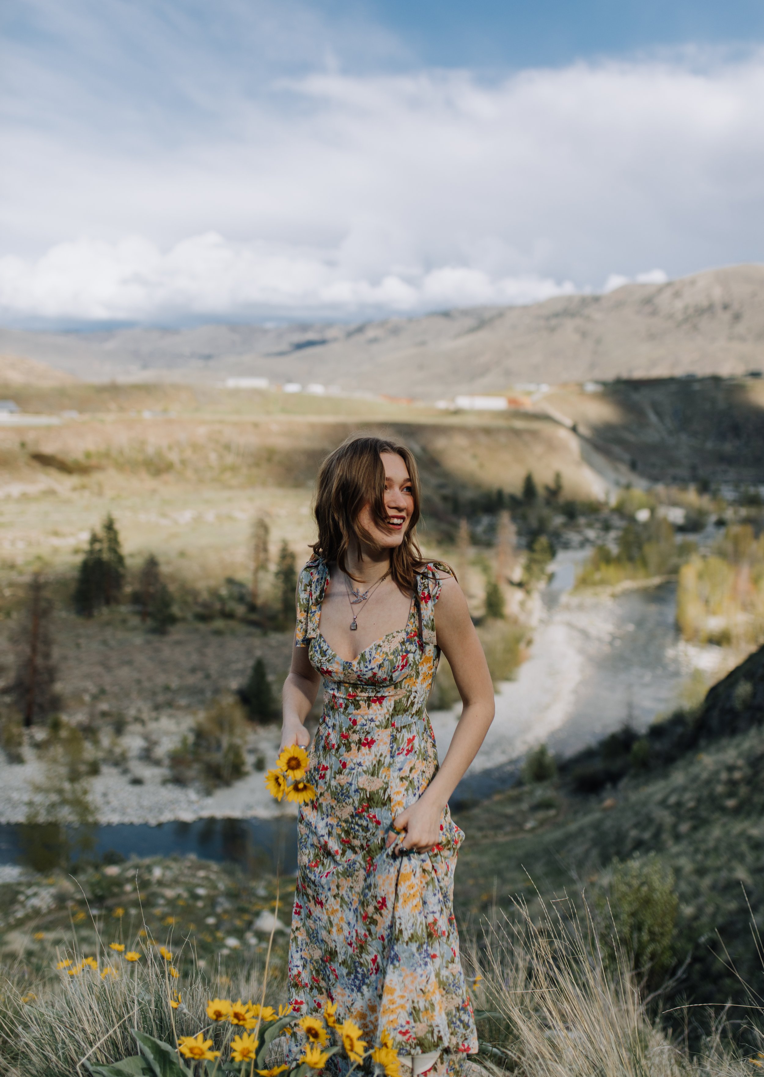 A young woman in a floral dress holding yellow flowers, smiling outdoors in a mountainous landscape with a river, trees, and cloudy sky in the background.