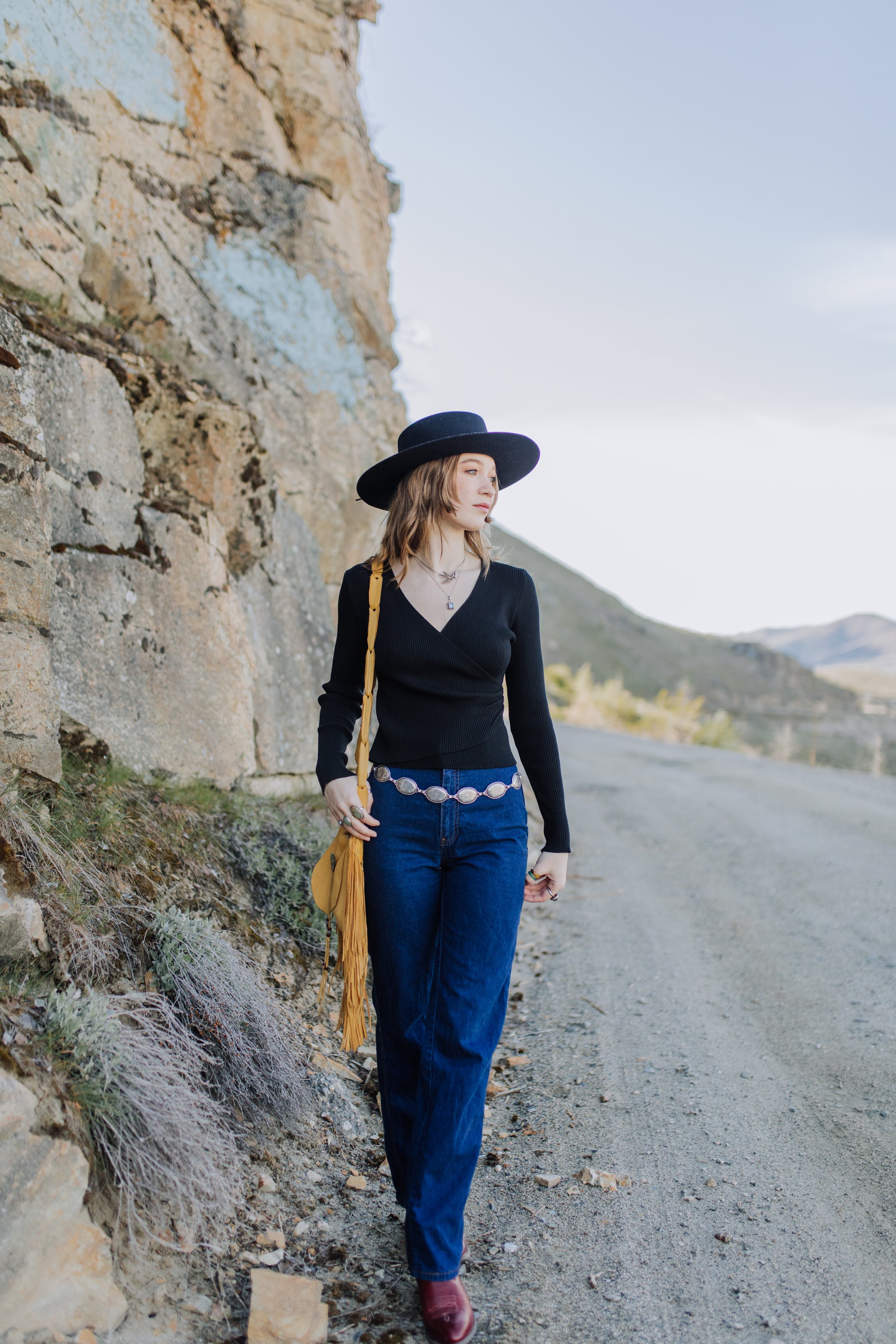 Woman wearing a black hat, black sweater, blue jeans, and maroon boots walking along a dirt road with a rocky hillside in the background.