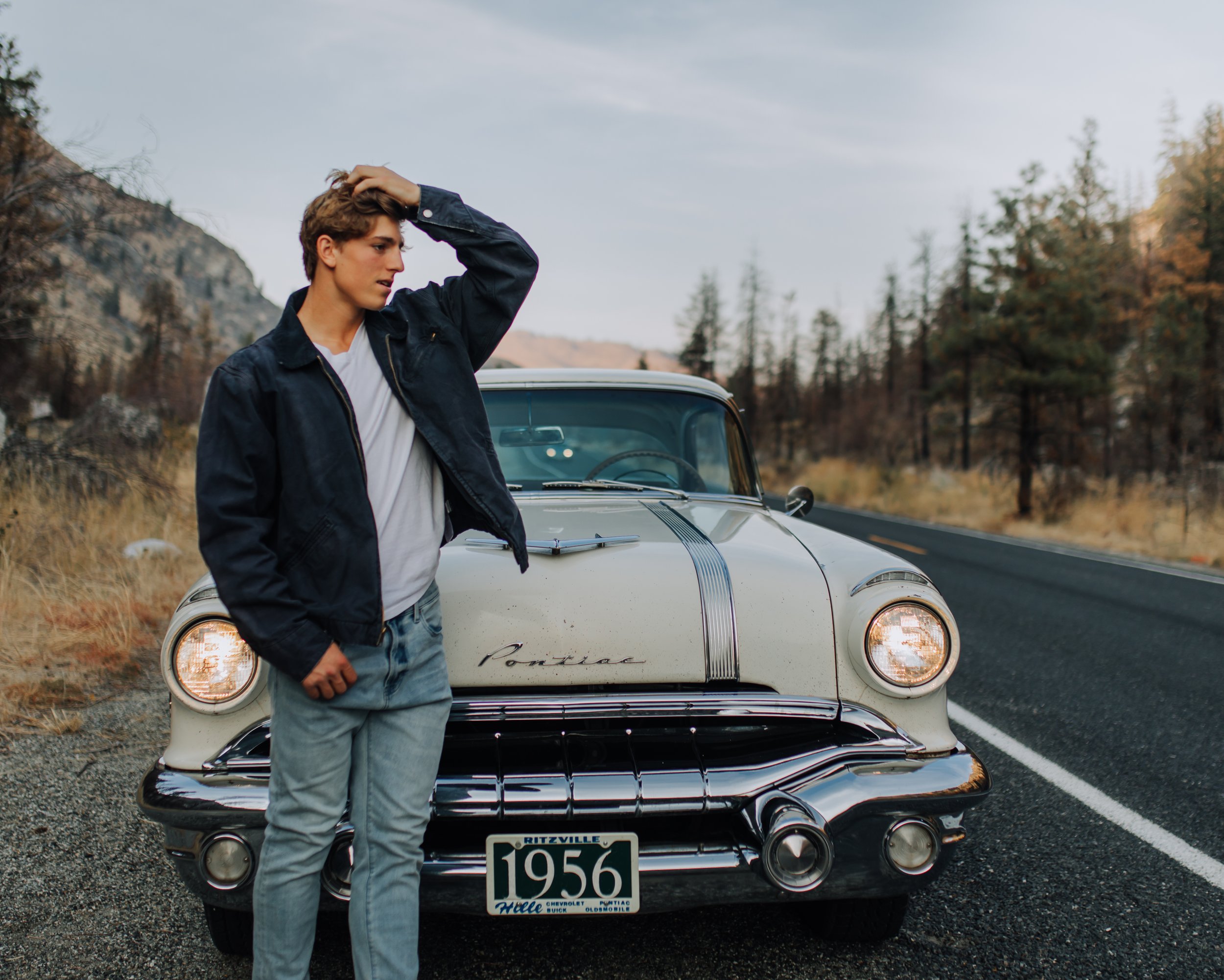 A young man leaning against a vintage white Pontiac car on the side of a rural road with autumn trees and mountains in the background.