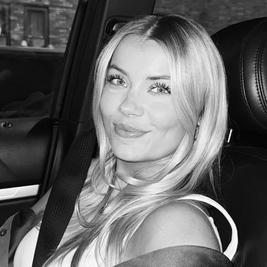 A woman with long blonde hair is sitting in a car and smiling at the camera.