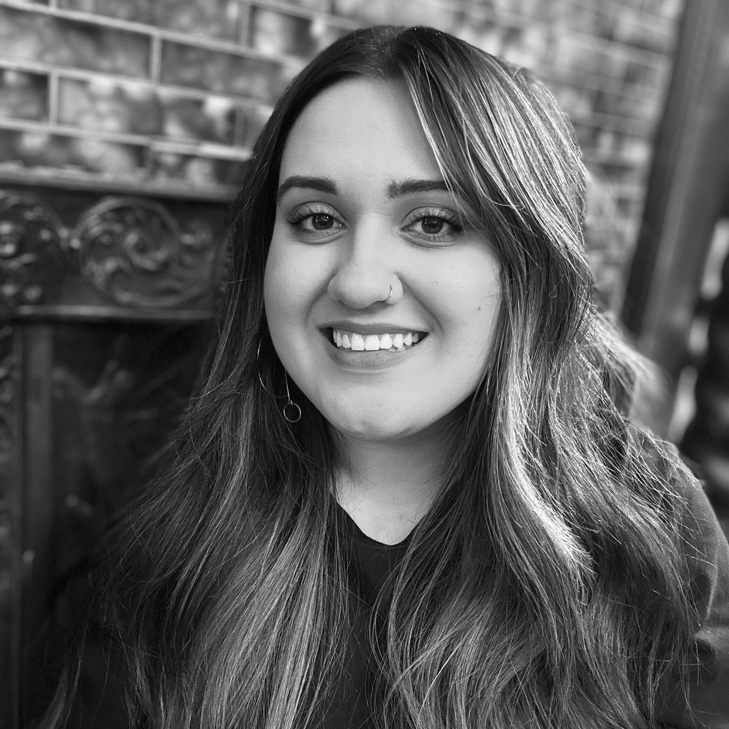 A smiling woman with long hair, wearing a nose ring, earrings, and a dark top, poses in front of a decorative metal and brick background.