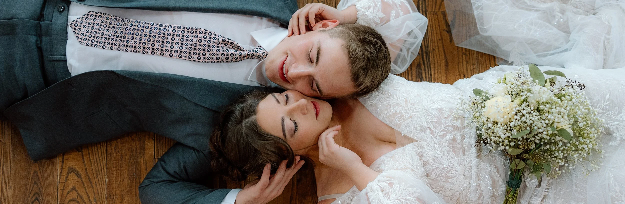 A newlywed couple lies on the wooden floor with their heads touching, eyes closed, smiling softly. The bride holds a bouquet of white flowers, and the groom wears a dark suit with a patterned tie. The scene captures a tender moment of happiness and love.