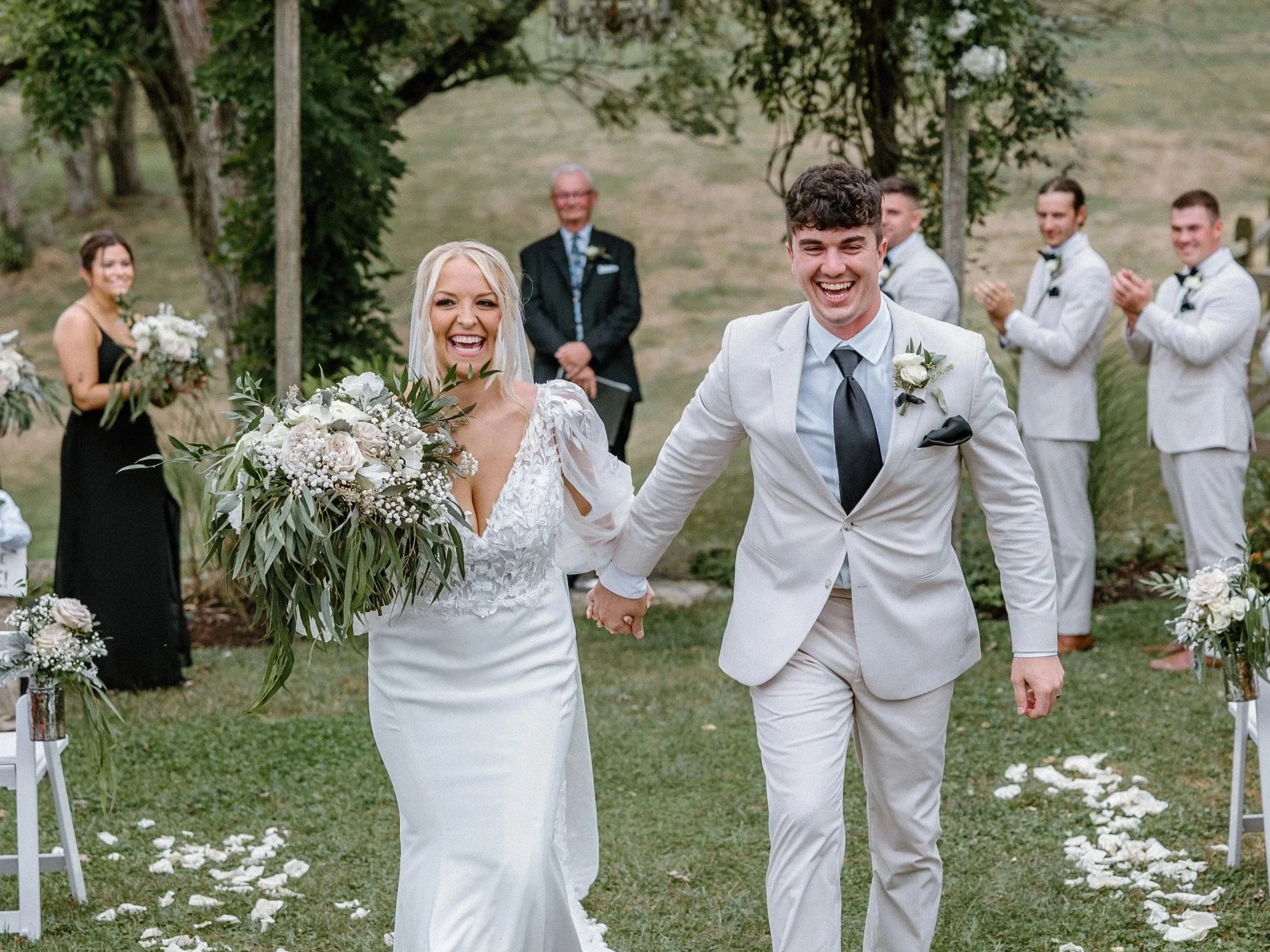 Happy bride and groom walking down an outdoor wedding aisle holding hands, surrounded by friends and family applauding, with a decorative floral arch in the background.
