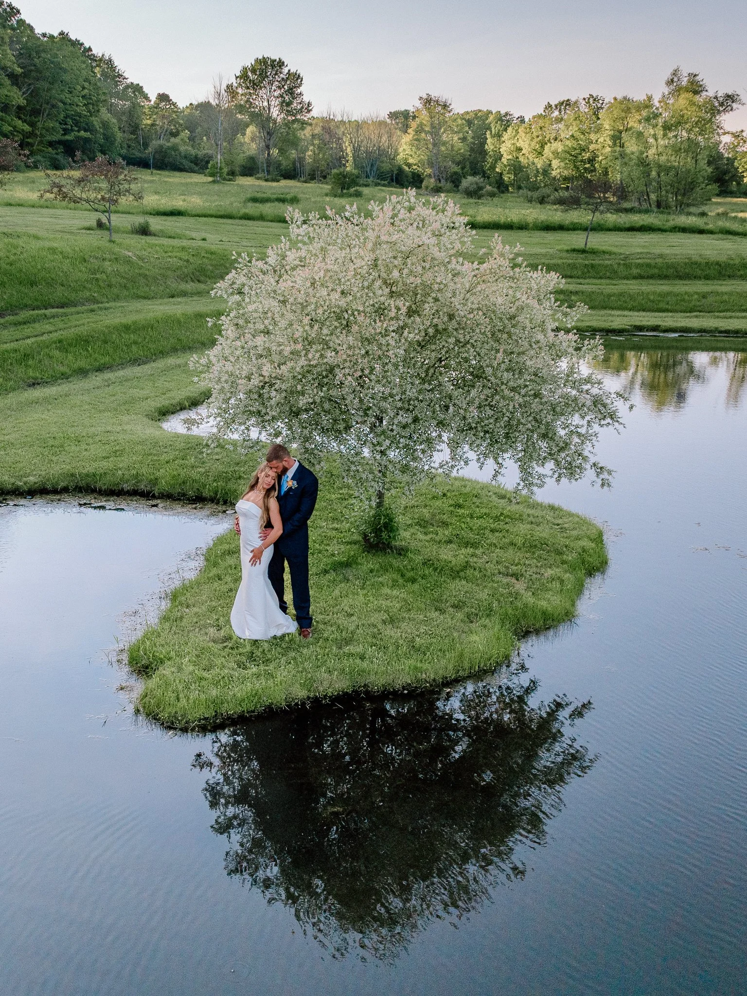 A bride and groom embracing on a small grassy island by a calm body of water, with a white flowering tree behind them and a green landscape with trees in the background.