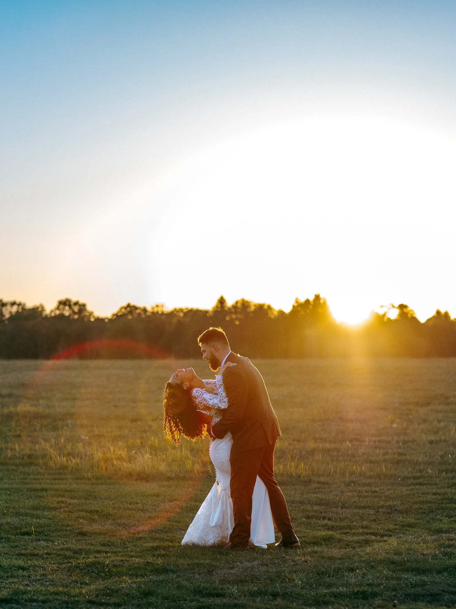 A bride and groom dancing outdoors at sunset in a field, with the groom dipping the bride.
