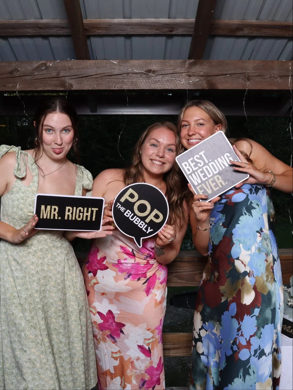 Three women posing with photo booth props at a wedding reception. The woman on the left is holding a sign that says "MR. RIGHT." The woman in the middle is holding a speech bubble that says "POP THE BUBBLY," and the woman on the right is holding a si