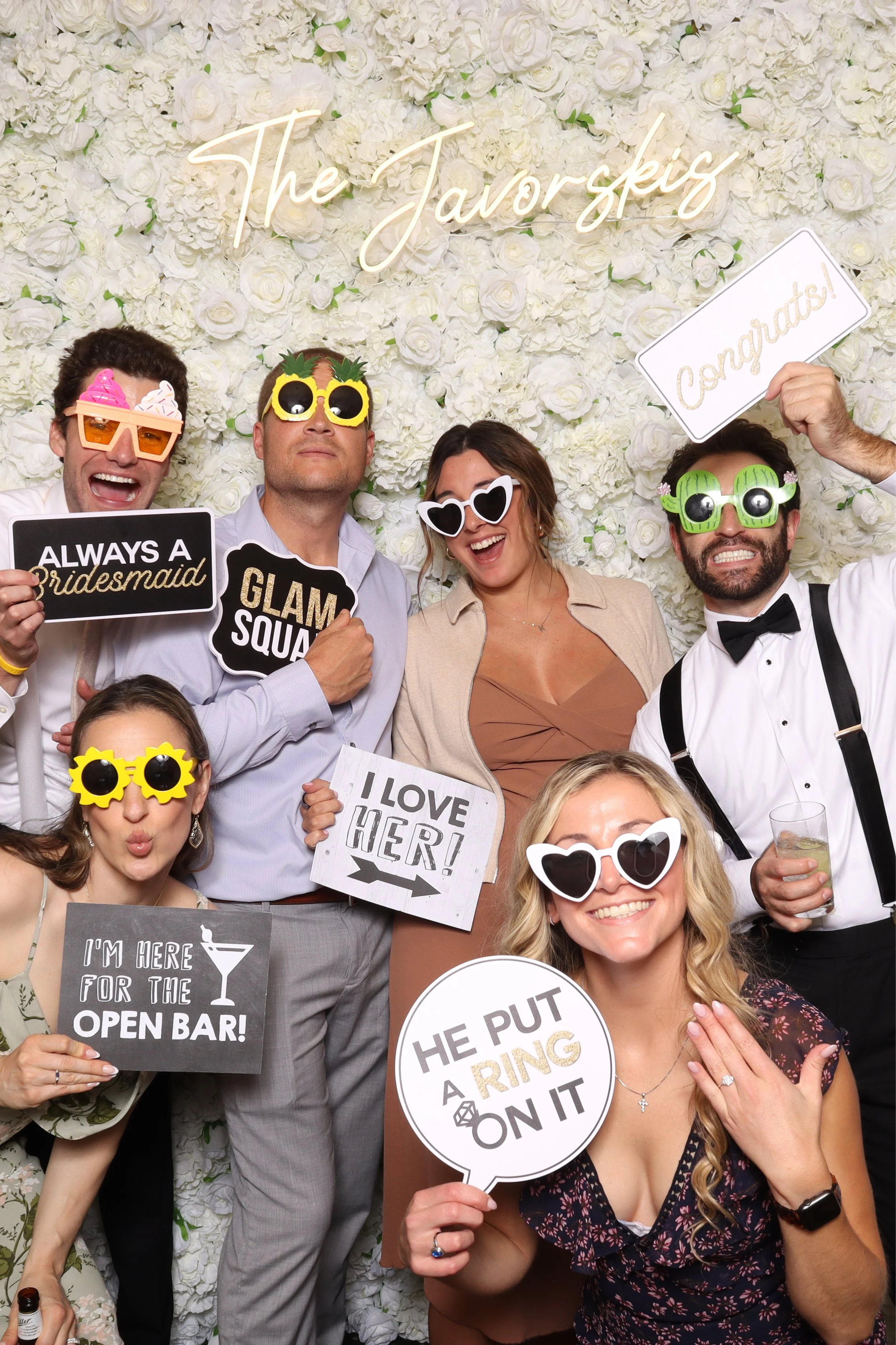 Group of people in party attire with photo props, posing in front of a floral wall with the words 'The Javorskis' in neon. They hold signs with humorous messages and wear costume glasses.