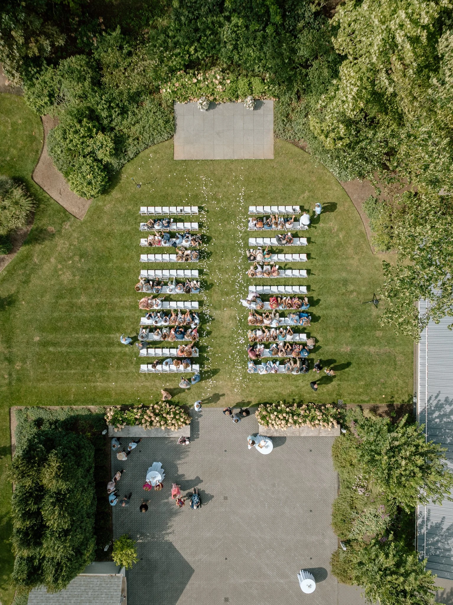 An aerial view of an outdoor wedding ceremony setup showing rows of white chairs on a grassy lawn, decorated with flower petals, with a small stage at the top and guests seated, surrounded by lush green trees.