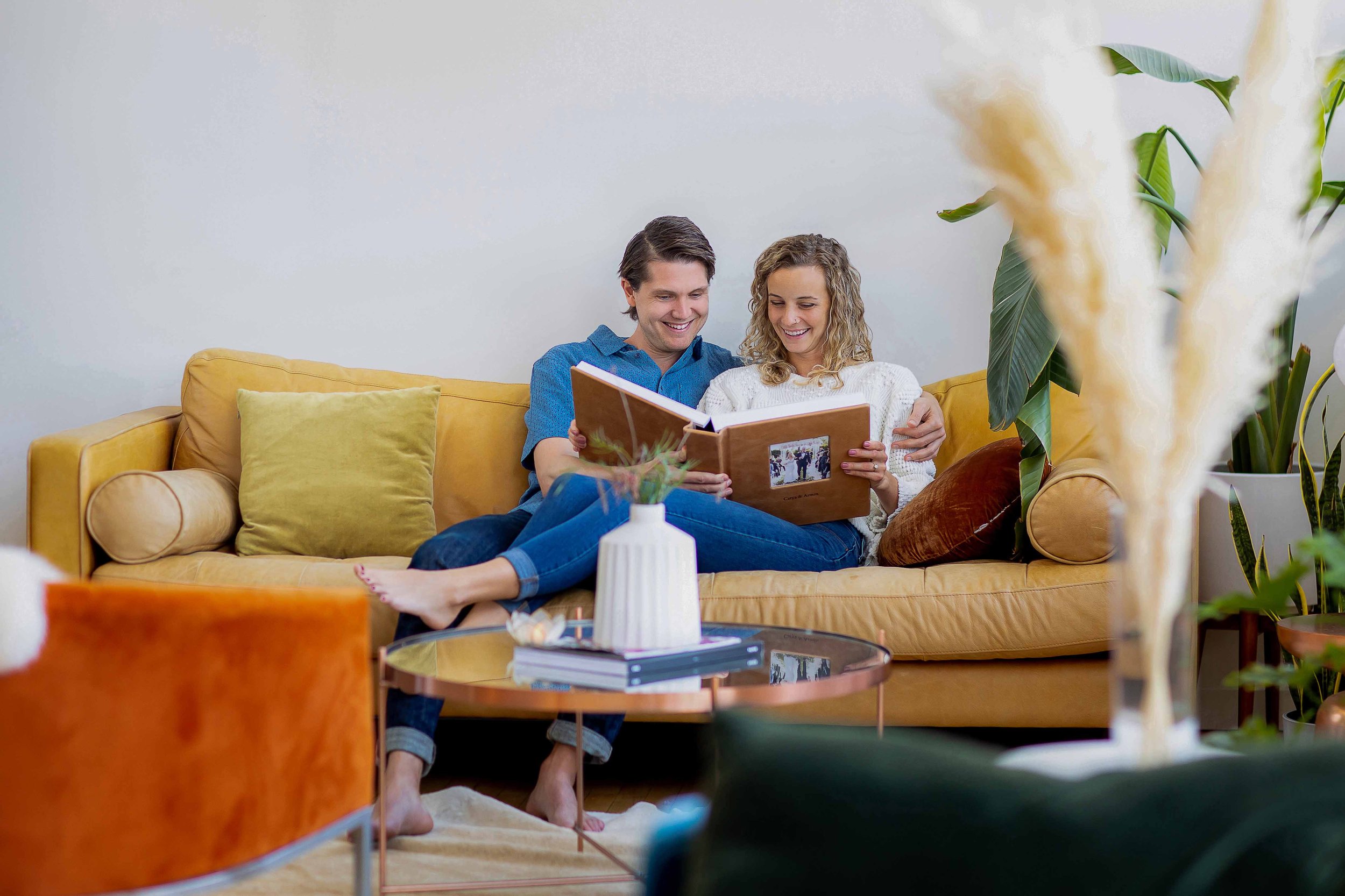 A happy couple sitting on a yellow sofa, looking at a photo album together, in a cozy living room with plants and warm lighting.