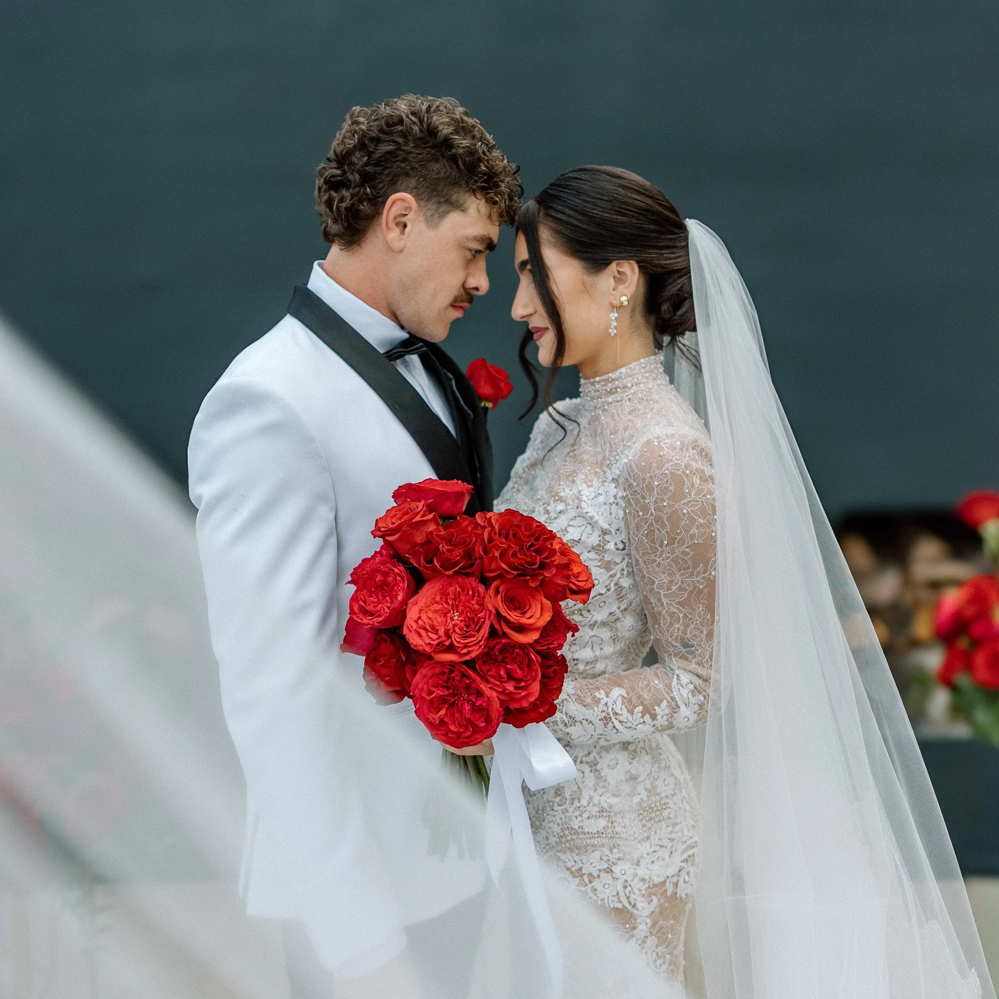 A bride and groom with foreheads touching, eyes closed, holding a bouquet of red roses, dressed in traditional wedding attire, against a dark gray background.