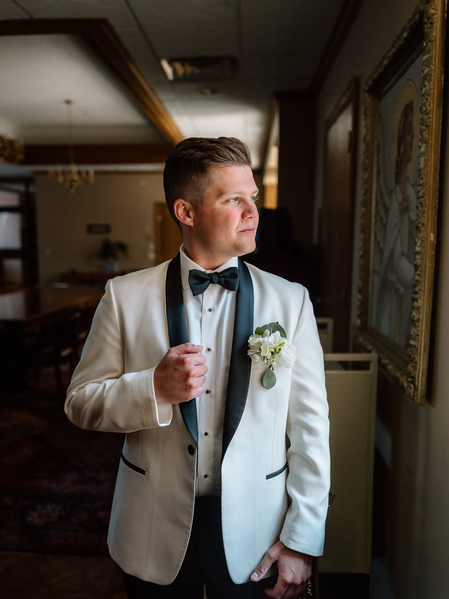 Man in a white tuxedo with black lapels and bow tie, holding a white flower boutonniere, looking sideways in a room with dark wood furniture and framed art.