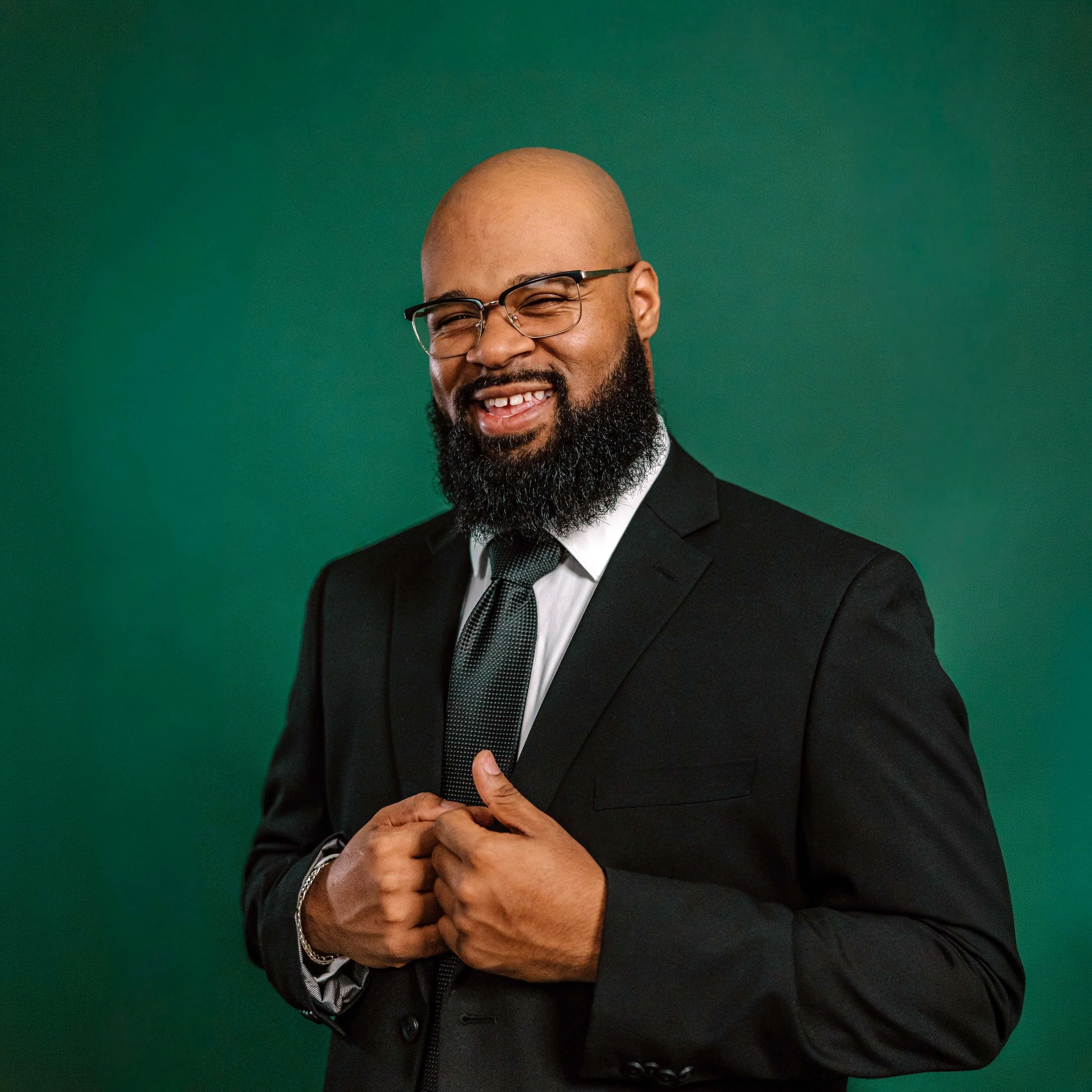 Close-up of a smiling man with glasses, bald head, and beard, wearing a black suit, white shirt, and black tie, against a plain green background.