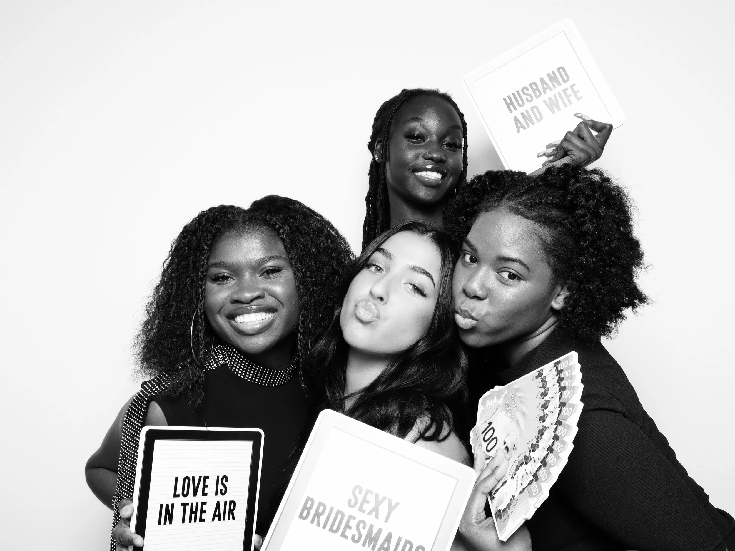 Four women smiling and posing for a photo in a photo booth. They are holding signs and props including a fan of dollar bills, a sign reading 'LOVE IS IN THE AIR', another sign reading 'SEXY BRIDESMAID', and a paddle with 'HUSBAND AND WIFE' written on