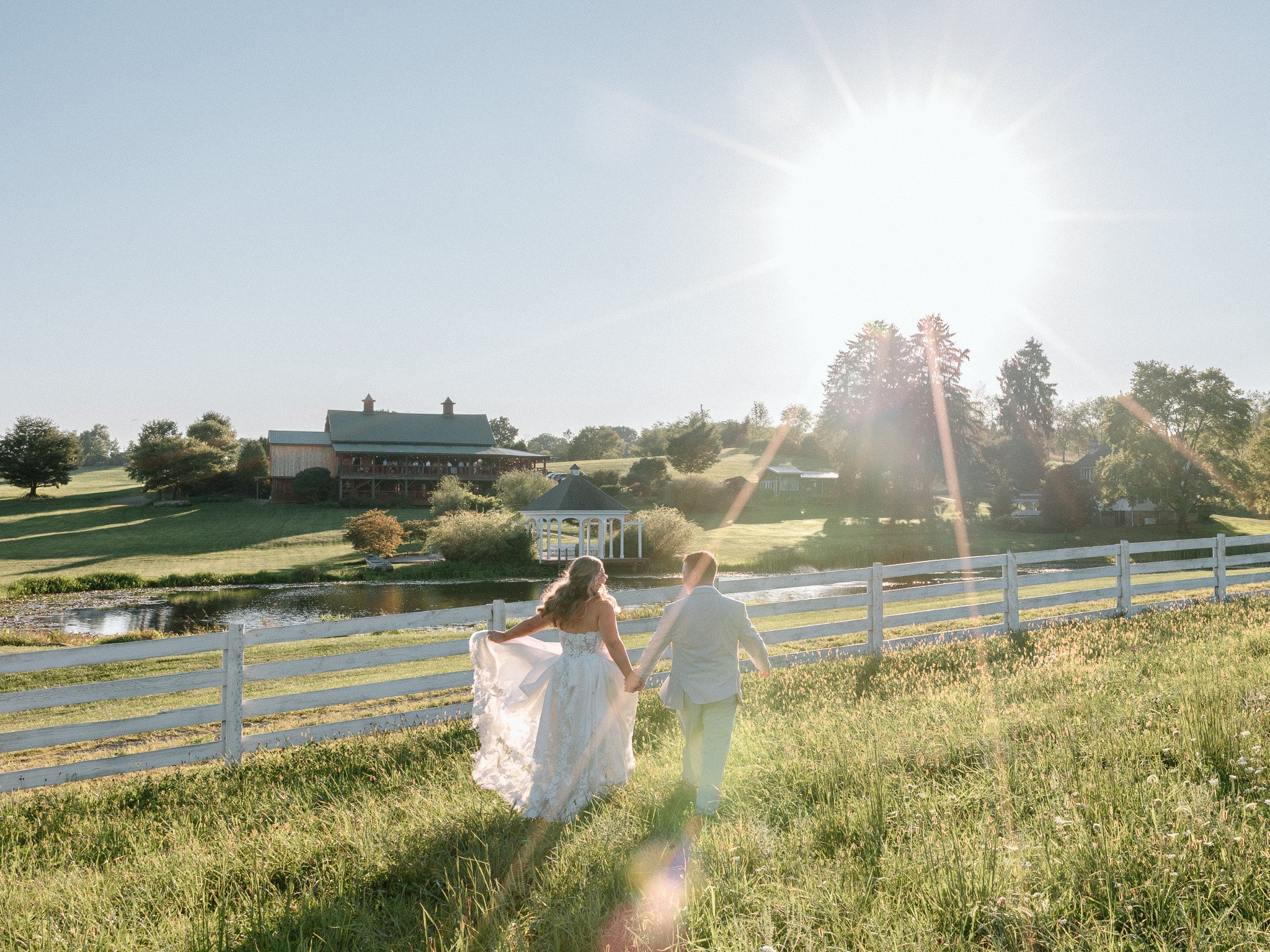 Bride and groom walking hand in hand into the sunset, celebrating outdoors in a field with a barn in the distance