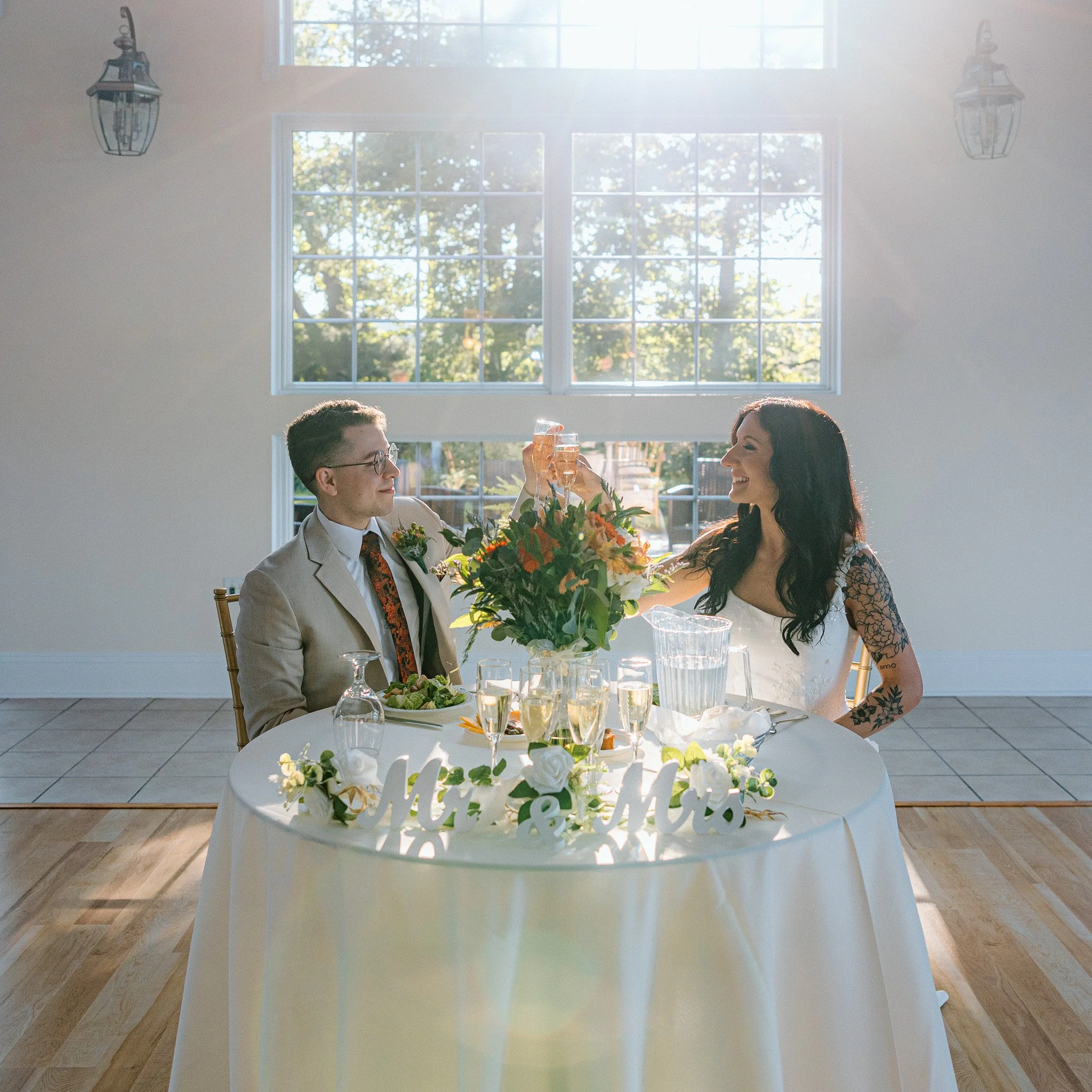 Two people, a man and a woman, are sitting at a table in a room with large windows, clinking glasses in a toast. The table is decorated with flowers and has several glasses of drinks on it. Sunlight streams through the windows, illuminating the room.