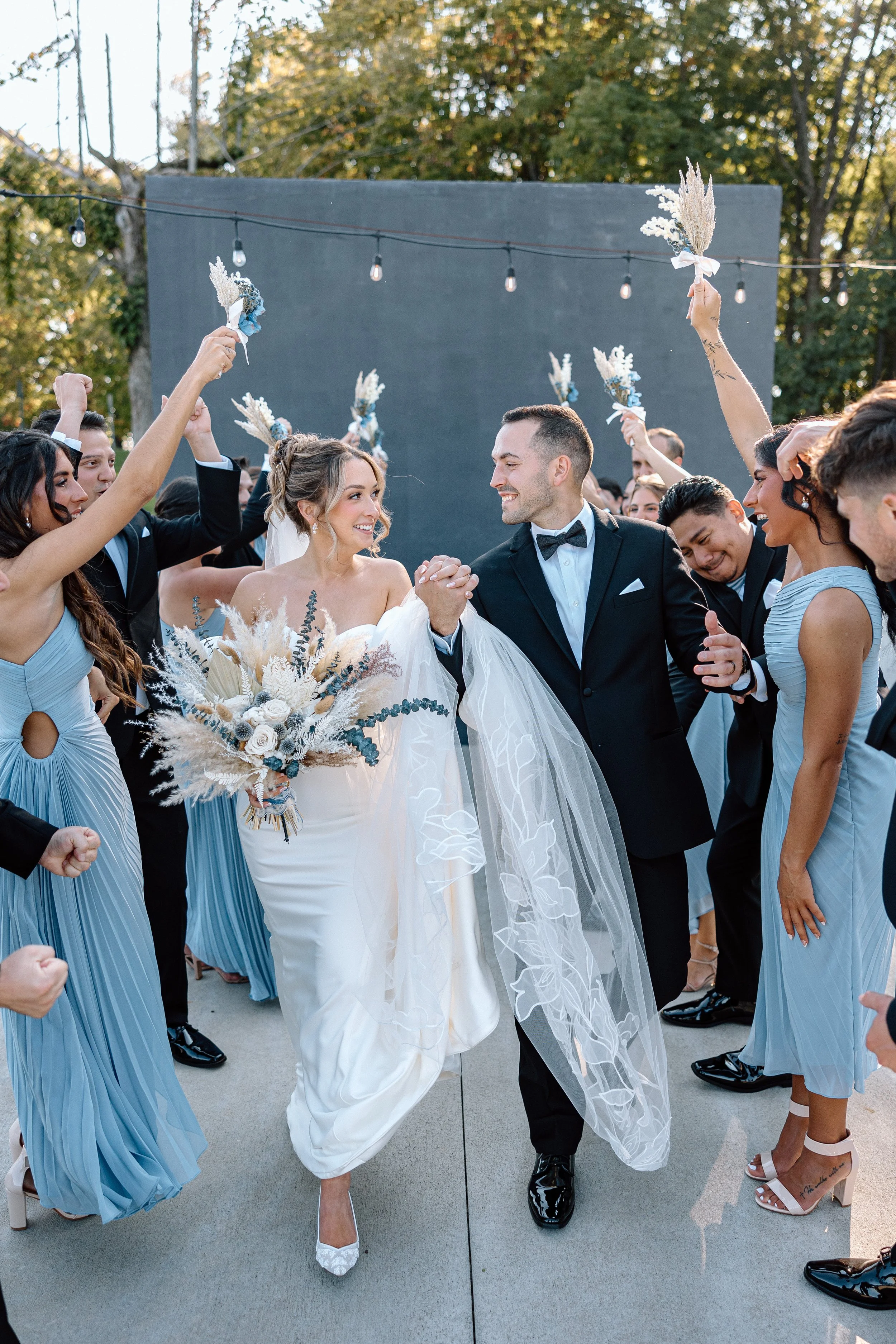 Bride and groom walking hand in hand, surrounded by wedding guests throwing flower bouquets and celebrating outdoors.