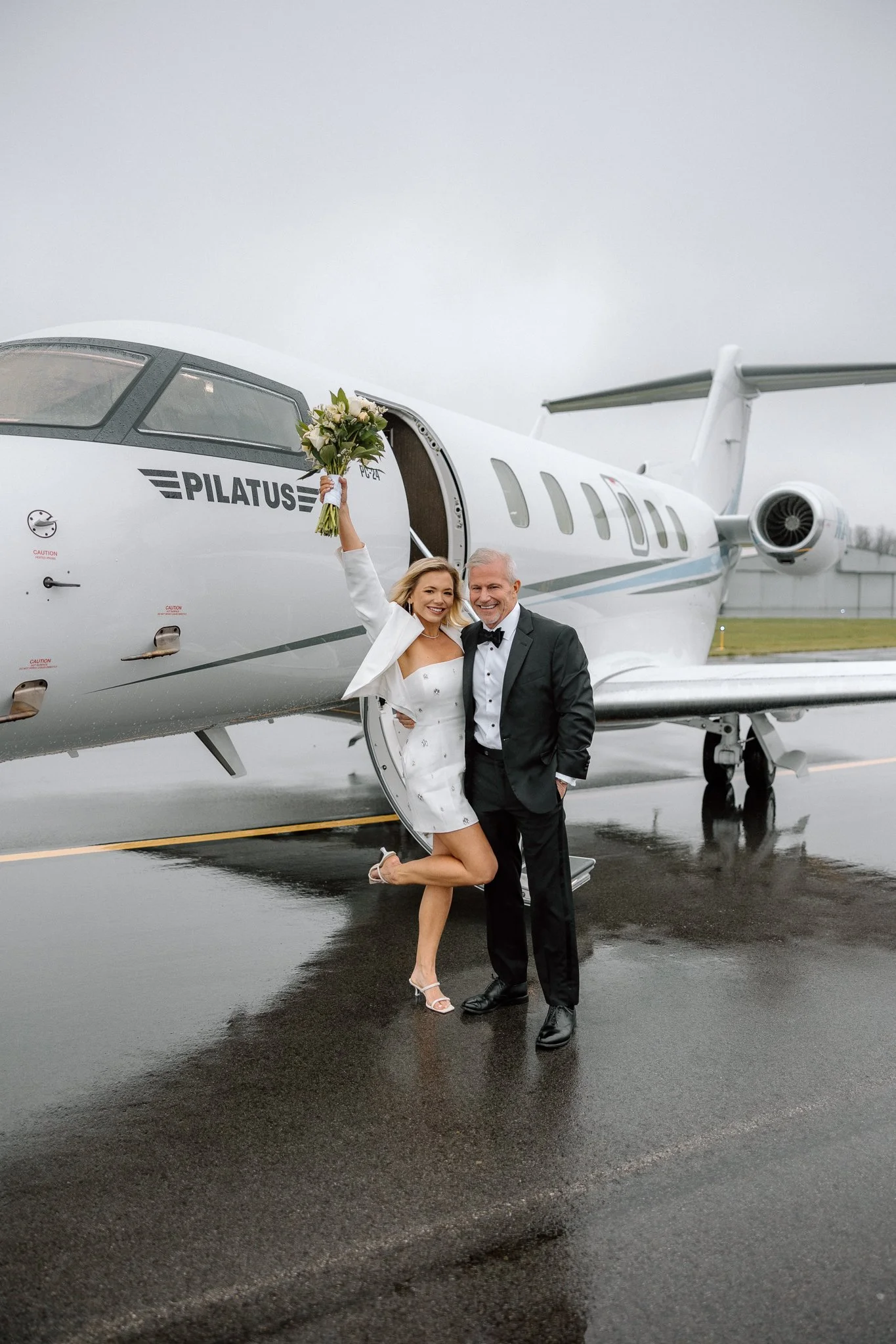 A woman in a white dress and high heels holding a bouquet of flowers, standing on her knee, next to a man in a tuxedo, both smiling, in front of a private jet on a wet tarmac.