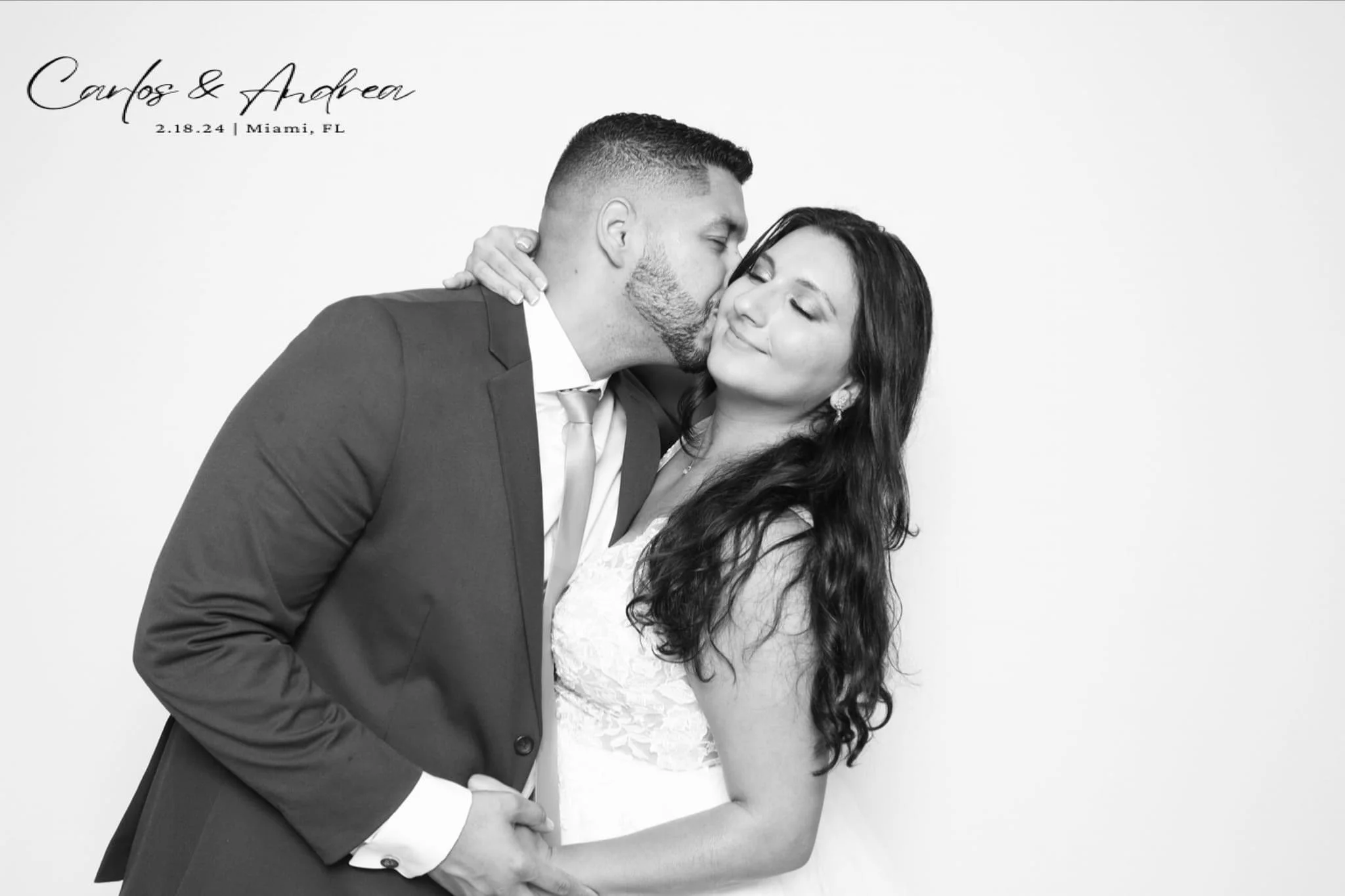 A black and white photo of a newlywed couple, with the groom kissing the bride on the cheek. The bride is smiling with her eyes closed, and they are holding hands. The top left corner has handwritten-style text that reads "Carlos & Andrea" with the d
